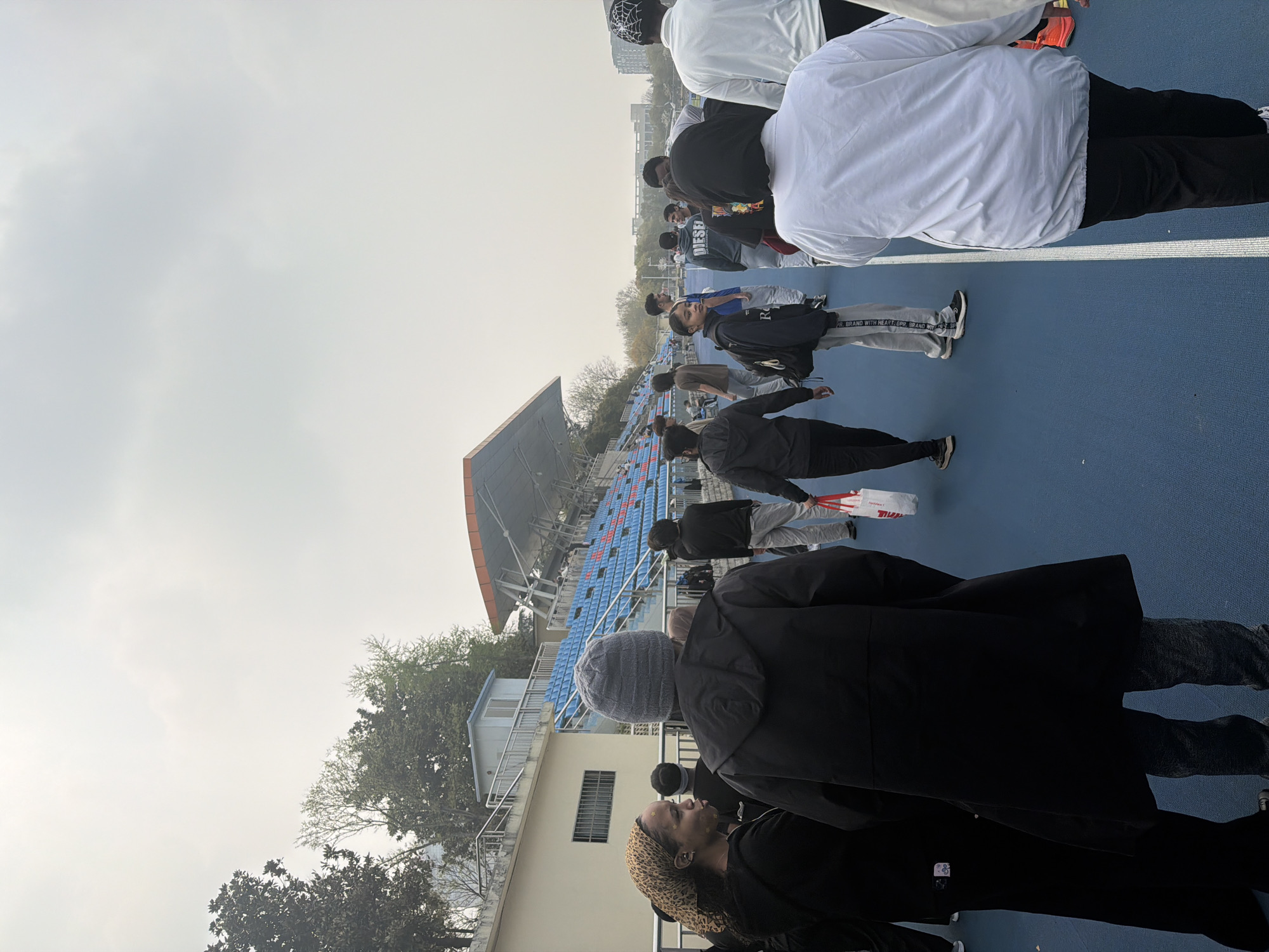 The image depicts a group of people standing on a blue track surface, likely a running track in a stadium. In the background are stadium seats, predominantly blue. Architectural features suggest the location is in the Jiangning District, China. The sky is overcast. The people are dressed in a variety of clothing, including jackets and casual wear. The composition suggests a moment before or after an event, with the individuals possibly waiting or leaving the stadium.