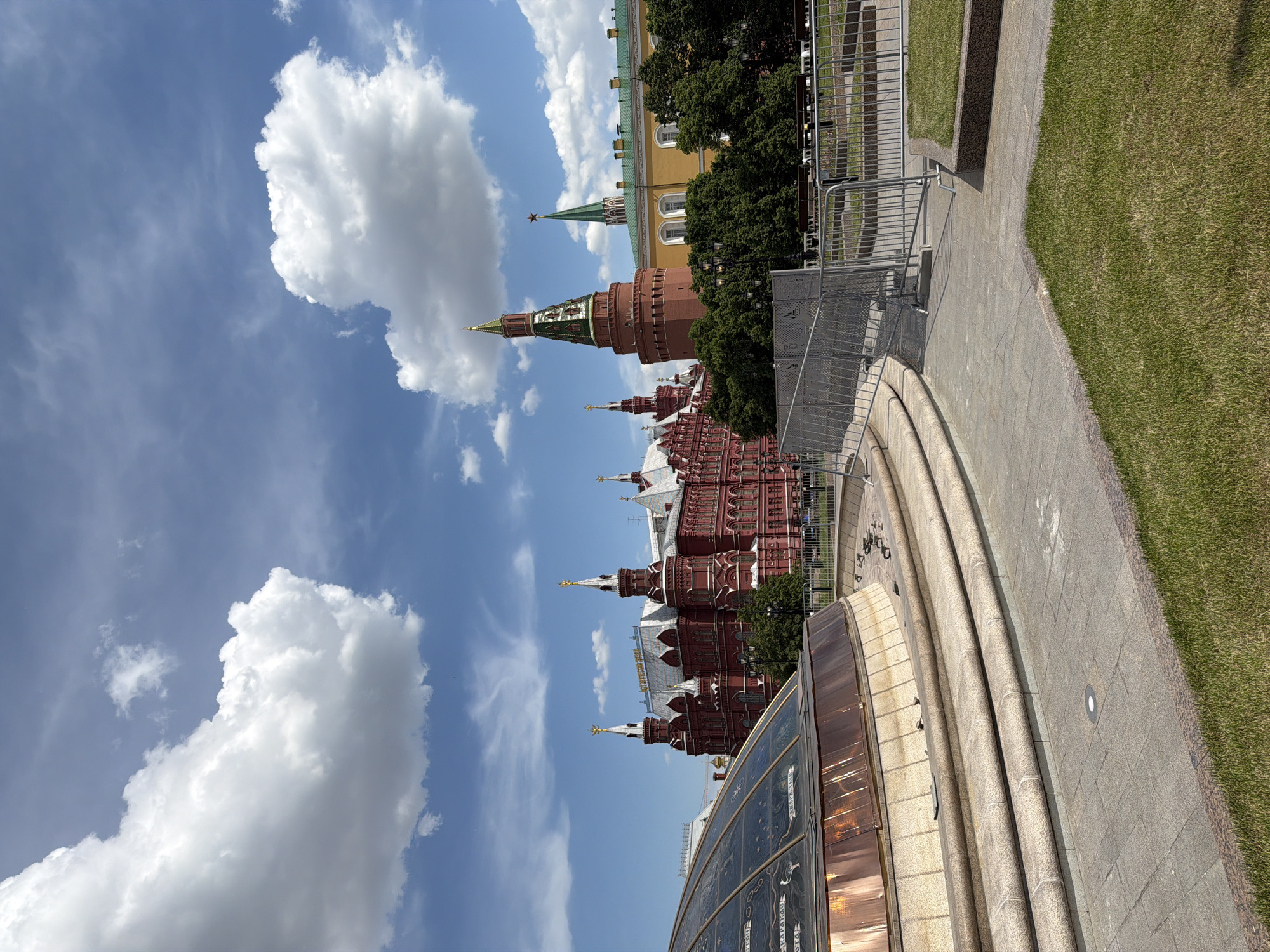 The image depicts a bright, sunny day in a historic urban setting in Moscow, Russia, showcasing prominent architectural landmarks under a blue sky.
In the foreground, a wide, paved pathway of grey tiles curves to the right, leading past a multi-tiered, stepped structure. A large, partially visible glass dome with a metal frame occupies the lower left corner, featuring faint, stylized etchings or decorative patterns that are not clearly legible. To the right of the pathway, a manicured lawn extends, separated by a low, curved border. Metal barriers or fences are visible along parts of the path and in the mid-ground.
The mid-ground is dominated by the majestic red brick facades of the State Historical Museum, characterized by intricate white decorative elements and multiple pointed spires, some topped with golden finials. Behind these, towards the left, a tall, round red brick tower with a green, conical spire topped by a red star (a distinctive Kremlin tower) rises against the sky. Further back and slightly to the right, a yellow building with a green roof, likely part of the Kremlin complex, can be seen peeking through dense green trees.
The sky is a vibrant blue, adorned with several large, fluffy white cumulus clouds, indicating fair weather. The strong illumination suggests it is daytime. There are no people visible, and no specific activities are taking place, presenting a serene, architectural vista. The combination of the Kremlin tower and the State Historical Museum clearly identifies the location as Moscow, Russia, likely in the vicinity of Red Square or Alexander Garden.