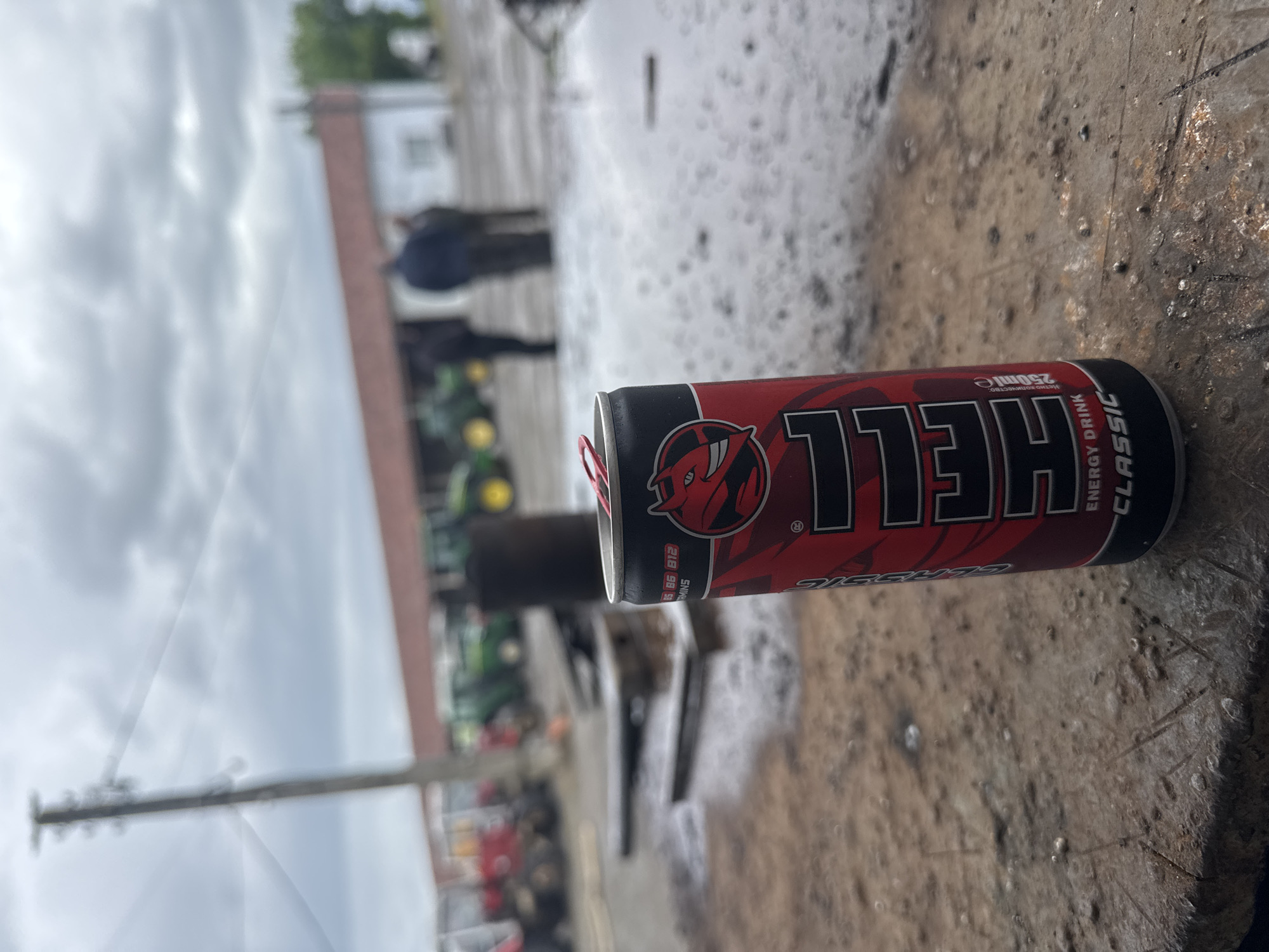 The image shows a can of "Hell" energy drink, Classic flavor, 250 ml, lying on a textured, weathered surface. In the background, blurred figures of people and tractors are visible, suggesting an agricultural or rural setting. A utility pole is present on the left side of the frame. The image was taken in Sarnevo, Bulgaria.