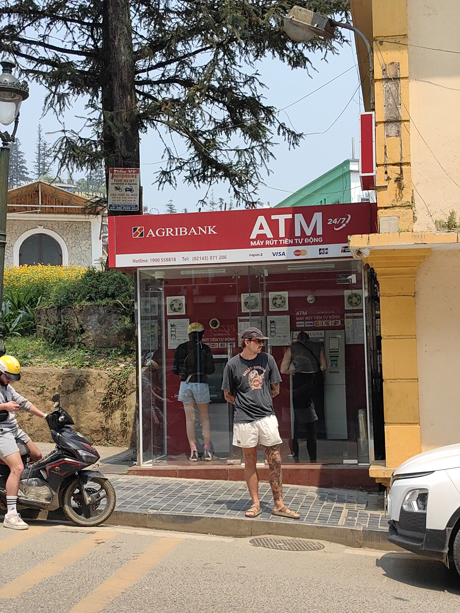 This image depicts an outdoor street scene in Sa Pa, Vietnam, centered around an Agribank ATM. The ATM booth features a prominent red facade with "AGRIBANK ATM 24/7" signage, including hotline and telephone numbers, and logos for Napas, Visa, Mastercard, and JCB. Inside the glass-fronted booth, two individuals are visible: one person in a dark jacket and light blue shorts stands at an ATM machine on the left, and another person in a dark vest and dark pants is at an ATM machine on the right.
On the tiled sidewalk in front of the ATM, a man stands wearing a black cap, sunglasses, a dark t-shirt, white shorts, and tan sandals, with extensive tattoos visible on his legs. He is looking towards the left. To his left, on the asphalt street which has faint yellow diagonal markings, a man wearing a yellow helmet, grey t-shirt, and grey shorts is seated on a black and red motorbike, appearing to look at a device in his hands. The front right corner of a white car is visible on the far right of the street.
The background includes a large tree with dark bark and sparse green foliage dominating the upper left, displaying a "MOTORBIKE FOR RENT" sign. A street lamp on a pole is visible on the far left. Behind the motorbike, a hillside covered with green foliage and yellow flowers leads to a building with a light-colored facade and a rounded archway. To the right, a yellowish-beige building facade with a column-like structure and an attached street light frame the ATM. The sky is clear and light blue.
