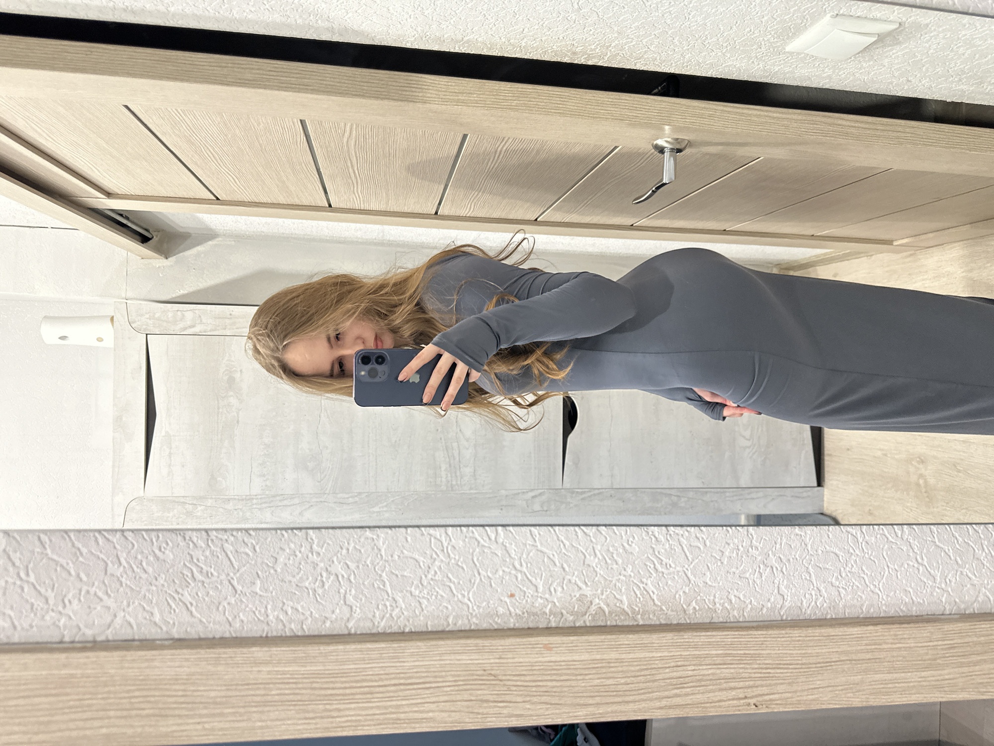 The image shows a woman taking a selfie in front of a mirror. She is wearing a long-sleeved, gray dress. Her hair is long and blonde. The background includes a wooden door, a textured white wall, and a shelf or vanity in front of the mirror. The location is unknown.