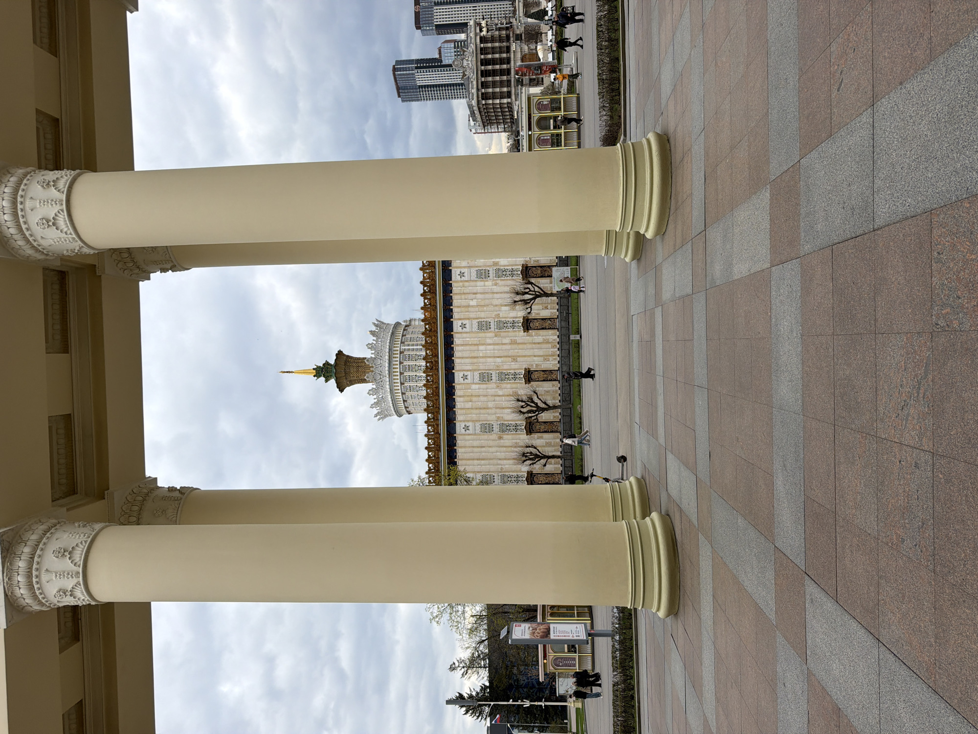 The image presents a grand architectural scene, likely captured from within a portico, overlooking a large public square or exhibition grounds in Moscow, Russia. Two prominent, light-colored classical columns with intricately carved capitals frame the view in the immediate foreground. The paved ground beneath is composed of square tiles, alternating in shades of reddish-brown and grey, arranged in a checkered pattern with distinct diagonal grey strips.
In the midground, a striking, ornate building with a golden spire and a multi-tiered, complex roof stands majestically. This structure, possibly an exhibition pavilion, features intricate architectural details. To its right, another building, adorned with decorative facades, is visible, fronted by several bare, stylized trees. Numerous people are seen walking casually throughout the paved areas, suggesting a public park or exhibition center environment. Near the right foreground column, a vertical sign is present, displaying text in Cyrillic, with "ШАУРМА" and "ФАЛАФЕЛЬ" being partially discernible, indicating food advertisements.
The background on the far left reveals several modern high-rise buildings, contrasting the older, ornate architecture and confirming an urban setting. The sky is overcast with scattered clouds, suggesting a daytime scene, possibly late morning or afternoon, under diffused light. The overall atmosphere conveys the scale and historical significance of the location, aligning with the characteristics of the All-Russian Exhibition Center (VDNH) in Moscow.