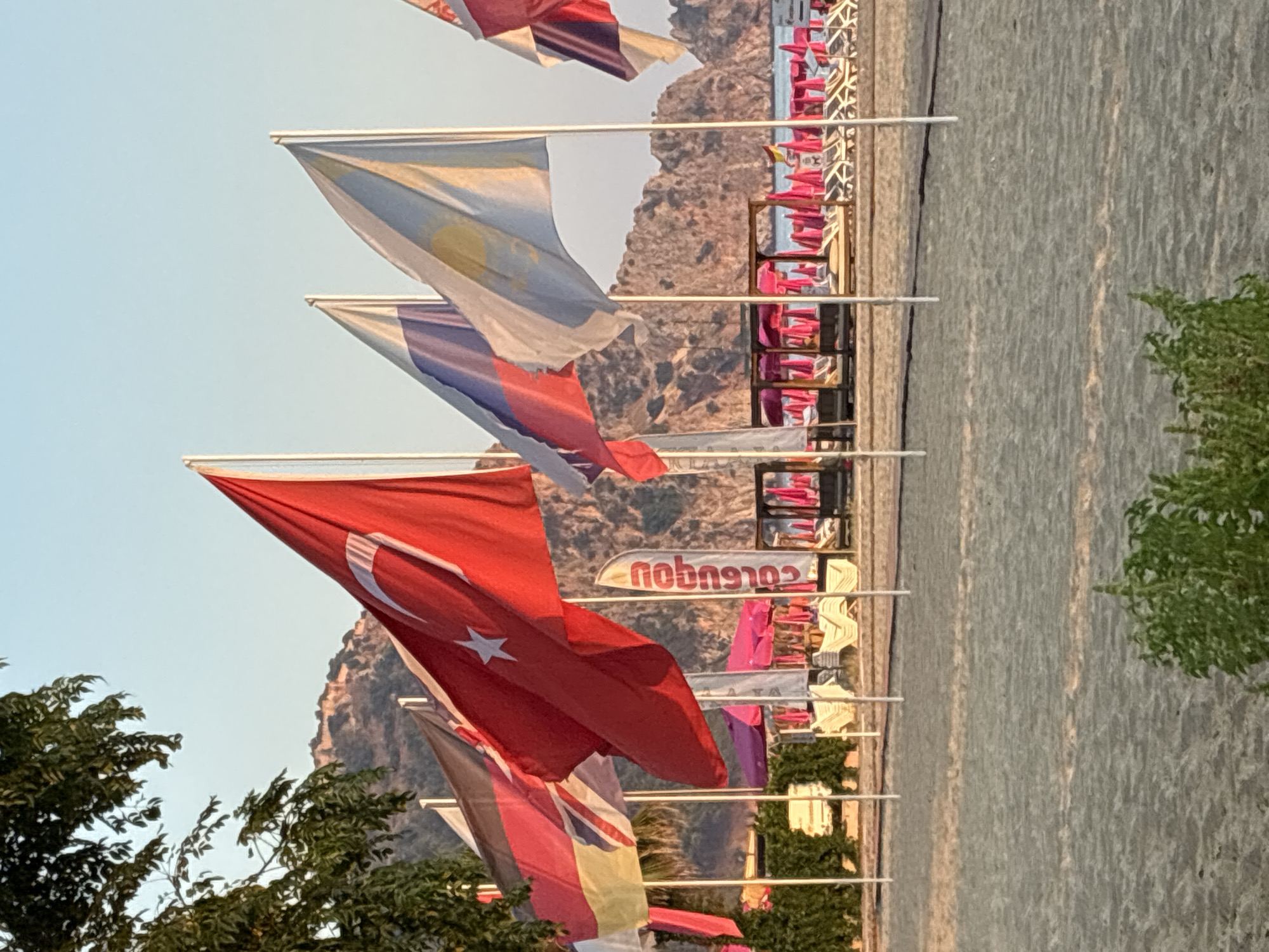 This image depicts a beach scene in Alanya, Turkey, with several national flags prominently displayed in the foreground. The flags include the Turkish flag, a German flag, a UK flag, a Russian flag, and a flag of Kazakhstan. Behind the flags, the beach is lined with rows of lounge chairs and umbrellas, indicating a tourist resort area. In the background, rugged hills and the clear sky suggest a warm, sunny day. The overall atmosphere is that of a popular tourist destination.
