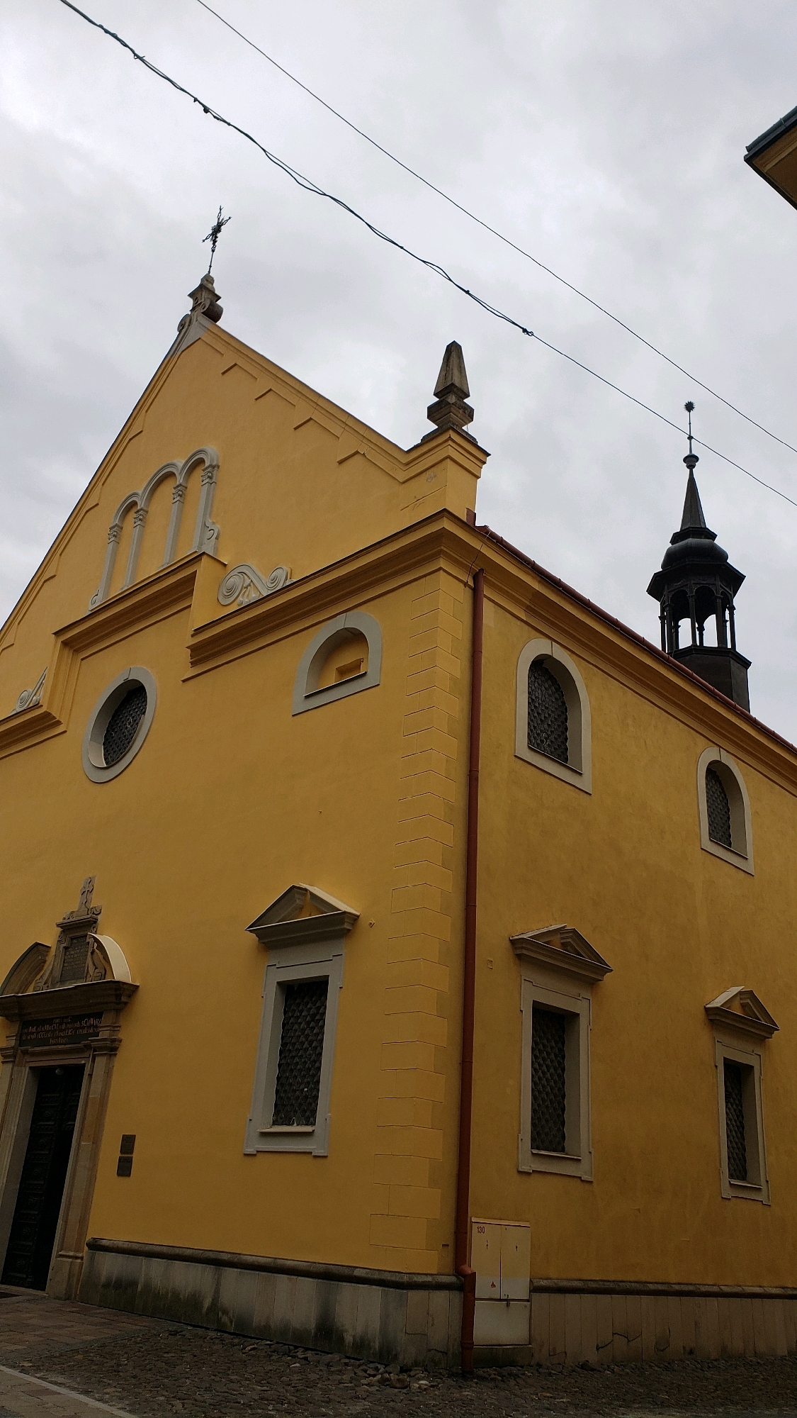 The image shows the exterior of a yellow building, likely a church, in Prešov, Slovakia. The building has a prominent entrance on the left with a dark door and an arched pediment above it. The facade is decorated with architectural details, including arched windows, a circular window, and a pitched roof topped with a cross. A black spire with an ornate top is visible on the right side of the building, suggesting it is a church tower. The sky is overcast, and power lines run across the upper part of the frame. The ground in front of the building is paved with cobblestones. The building appears to be well-maintained and is situated in an urban environment.