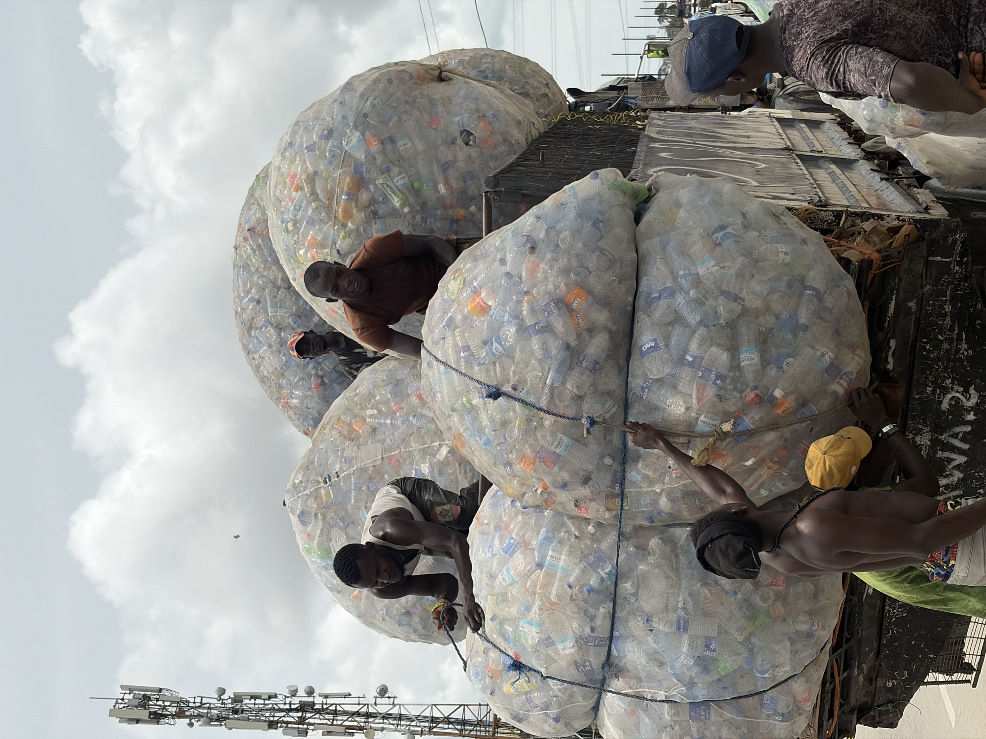 In Igboefon, Nigeria, a group of at least six men are working around a flatbed truck heavily laden with enormous, netted bags filled with plastic bottles. One man, wearing a brown shirt, stands atop the load. Another man, wearing a white shirt and a black tank top, works near the bottom of the pile. A third man is shown shirtless, wearing a yellow cap, while securing one of the bags. A radio tower is visible in the lower left corner against a cloudy sky. Other men are seen standing on the bed of the truck. They appear to be securing the bags with ropes, which encircle each netted bag. The focus is on the labor-intensive process of handling the collected bottles.