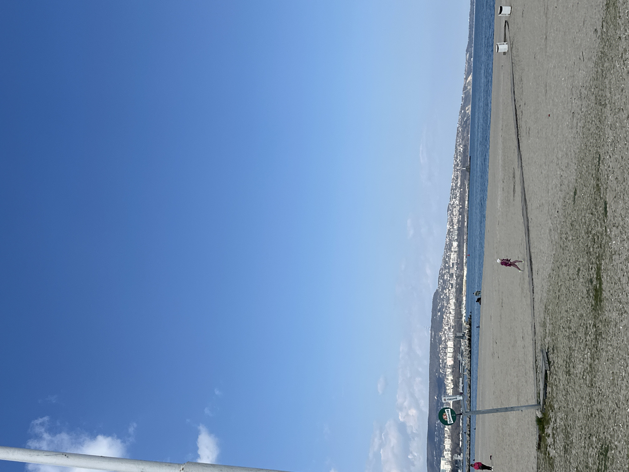The image shows a beach scene in Varna, Bulgaria. The sky is mostly clear and blue. To the right is the beach, with sand, water, and a city skyline visible in the distance. A person in pink clothing is walking on the beach. A sign is visible in the foreground near the beach. Another person in pink clothing is standing near the sign. The image is rotated counterclockwise.