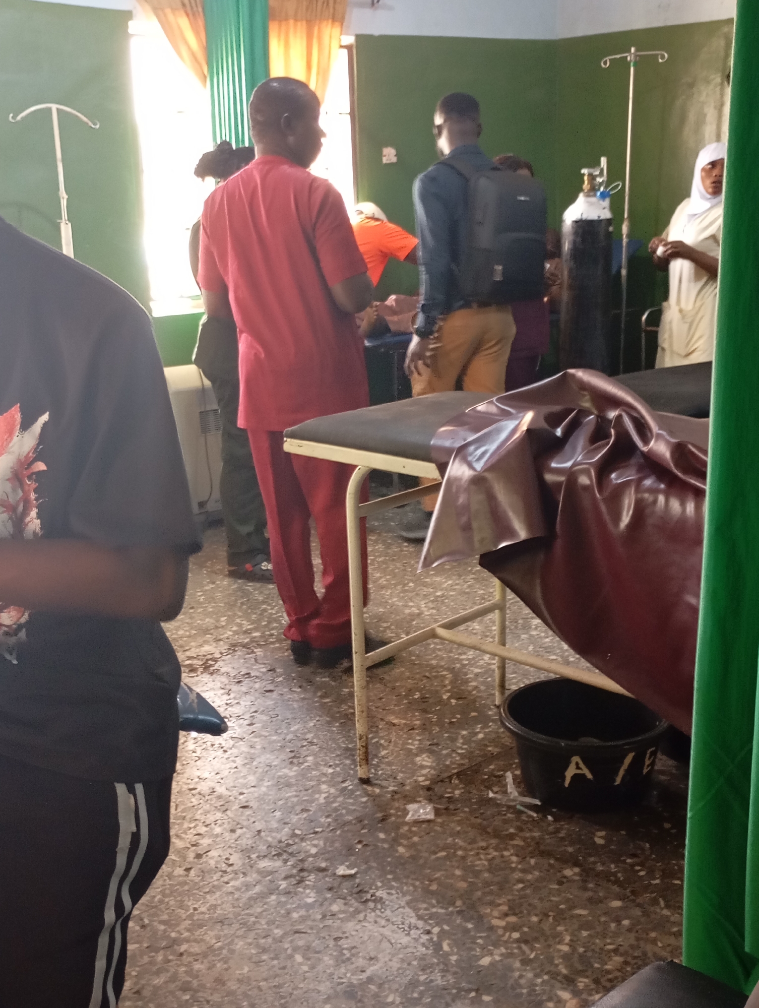 The image captures an indoor scene within a medical facility, likely a hospital ward or clinic in Jos, Nigeria. Several individuals are present, suggesting ongoing activities related to patient care.
In the foreground, on the left, a person is partially visible wearing a black t-shirt and track pants with grey stripes. A medical bed or stretcher, covered with a shiny maroon vinyl-like material, occupies the center-right foreground. Beneath this bed, on the dirty, speckled floor, a black bucket is visible with the letters "A/F" painted in white. A small piece of white plastic, possibly medical packaging, lies on the floor near the bucket.
In the mid-ground, a person in a red uniform, indicative of medical staff, stands facing away from the camera. Another individual, wearing a dark blue shirt, khaki pants, and a black backpack, also faces away, positioned near two large oxygen cylinders and an IV pole. To the far right, a woman in a light-colored headscarf and long garment stands, looking towards the right side of the frame. A glimpse of another person in an orange shirt is seen behind the person in the red uniform. An additional IV pole stands near a bright window on the far left, through which daylight streams, suggesting it is daytime. The walls are green, and the windows are adorned with orange and green curtains.
The scene depicts an active, functional medical environment, possibly a busy or resource-constrained facility, given the number of people and the state of the floor.