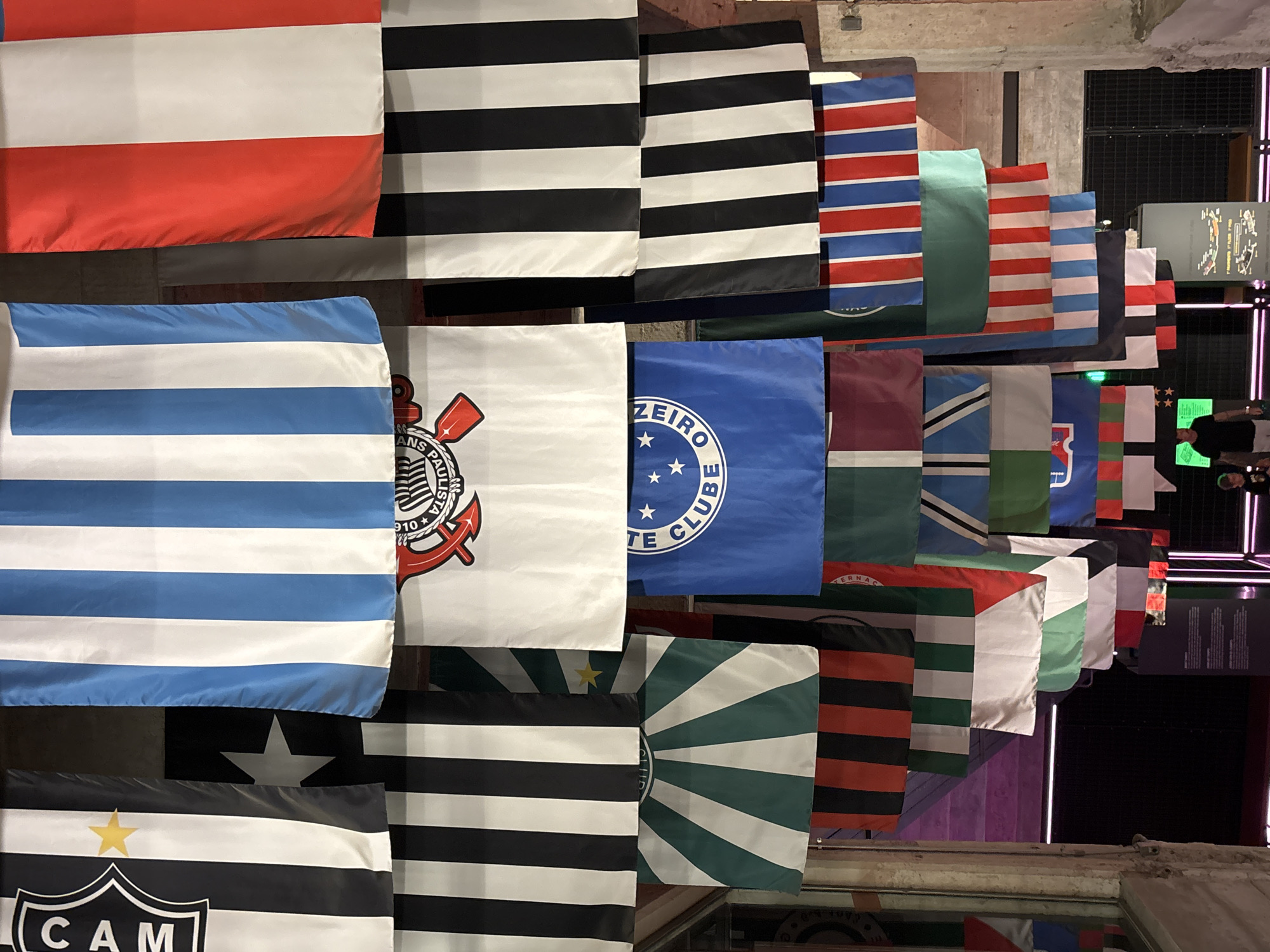The image shows a display of flags, likely inside a building. The flags represent various Brazilian football clubs, identifiable by their distinct colors and logos. Some recognizable clubs include Corinthians, Cruzeiro, and Atlético Mineiro. The flags are hung in rows, creating a layered effect. In the background, there are hints of an exhibition space with lighting and display panels. Based on the football flags and architectural style, the location is likely within the city of São Paulo, Brazil. Two people are visible in the background, near what appears to be a reflective surface.