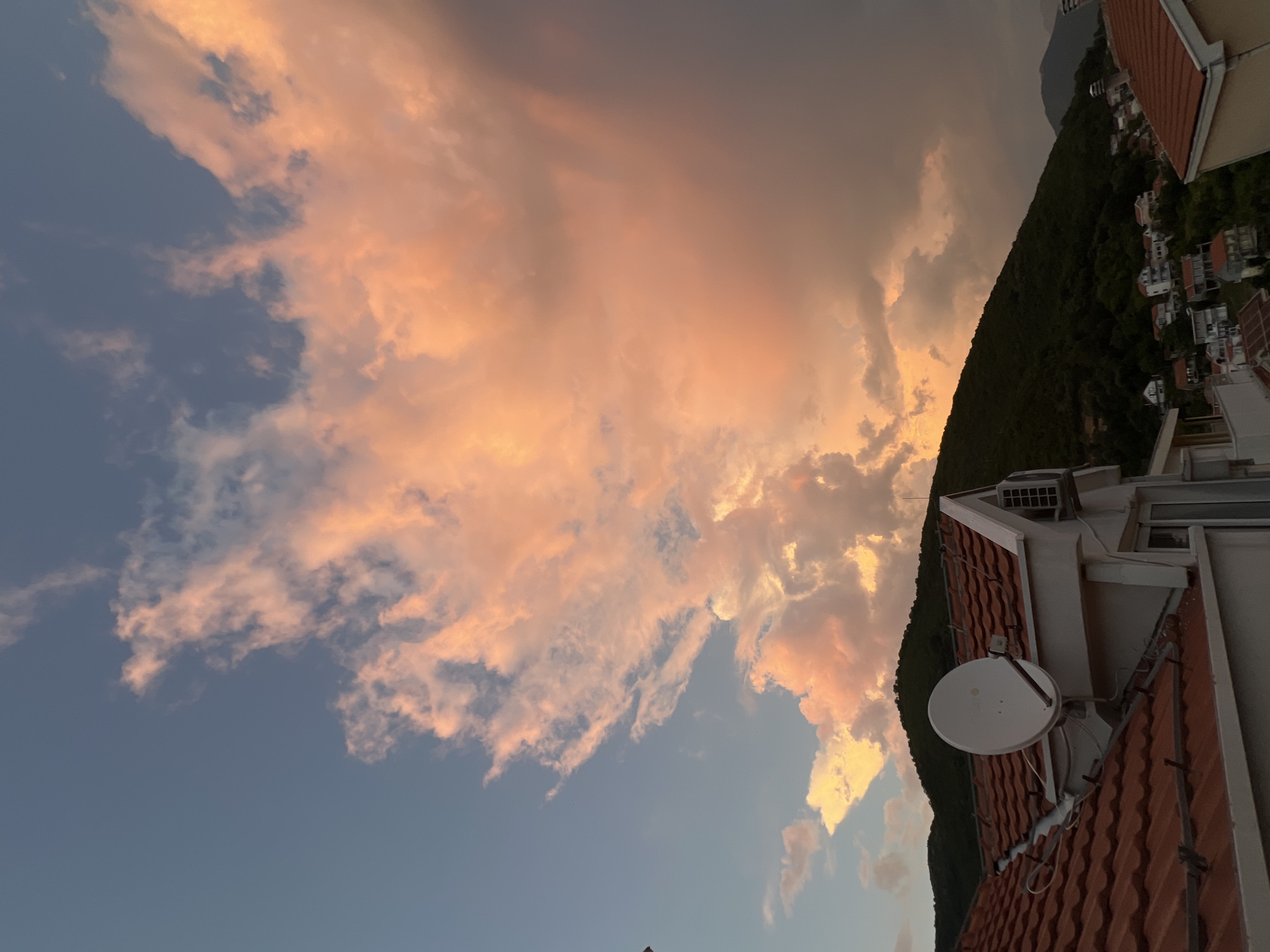 This is a vertical, wide shot of a sunset over Baošići, Montenegro. The sky is filled with dramatic clouds, tinged with orange and pink from the setting sun. In the foreground, a terracotta-tiled roof is visible, with a white satellite dish mounted on it. Behind the roof, a steep, dark green hillside is covered in trees, with scattered white houses nestled into its slopes. The overall mood is peaceful and serene, with the vibrant colors of the sunset creating a beautiful spectacle. The time of day is sunset, as indicated by the warm, colorful light illuminating the clouds. There is no visible text in the image.