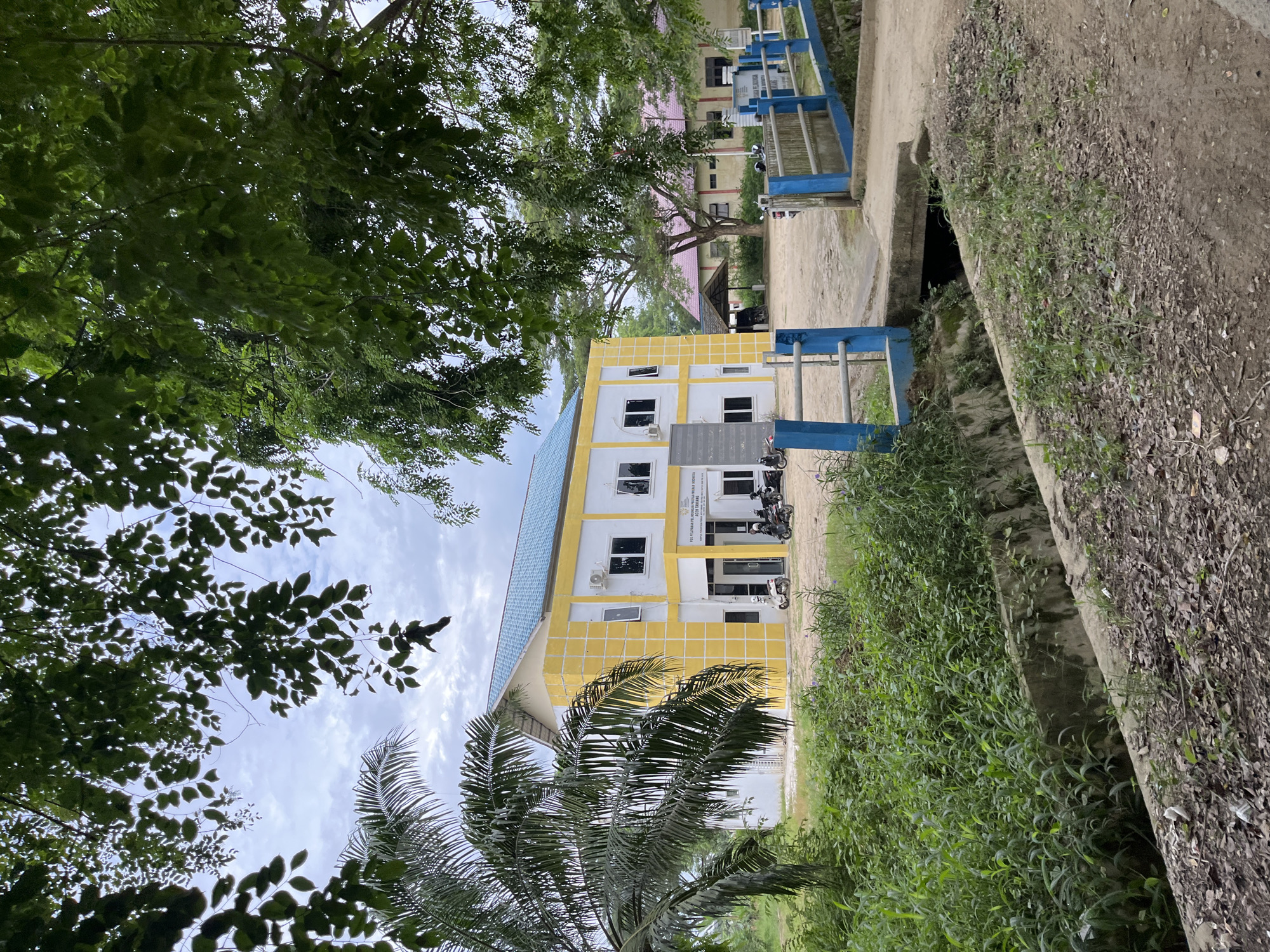 This is a rotated, outdoor, eye-level, medium shot of a building and its surroundings in Bundar, Indonesia. The building is painted yellow with white trim and has a blue tiled roof. It has multiple windows and air conditioning units. In front of the building, there are several motorcycles parked. A blue and white railing runs along the side of the building and a dirt path. To the right of the building, there is a concrete channel with overgrown green vegetation and dry leaves. The sky is cloudy, suggesting it might be an overcast day. The overall scene appears to be that of a public building or institution, possibly a school or administrative office. The time of day is indeterminate due to the lighting, but the cloudy sky suggests it is daytime. The image lacks any discernible emotional cues, but the presence of the building and motorcycles implies a functional location. Visible text on a sign above the entrance reads "PELATIHAN PEKERJA SOSIAL INDONESIA" and "BALAI PELATIHAN PEKERJA SOSIAL", indicating it is a social worker training facility.