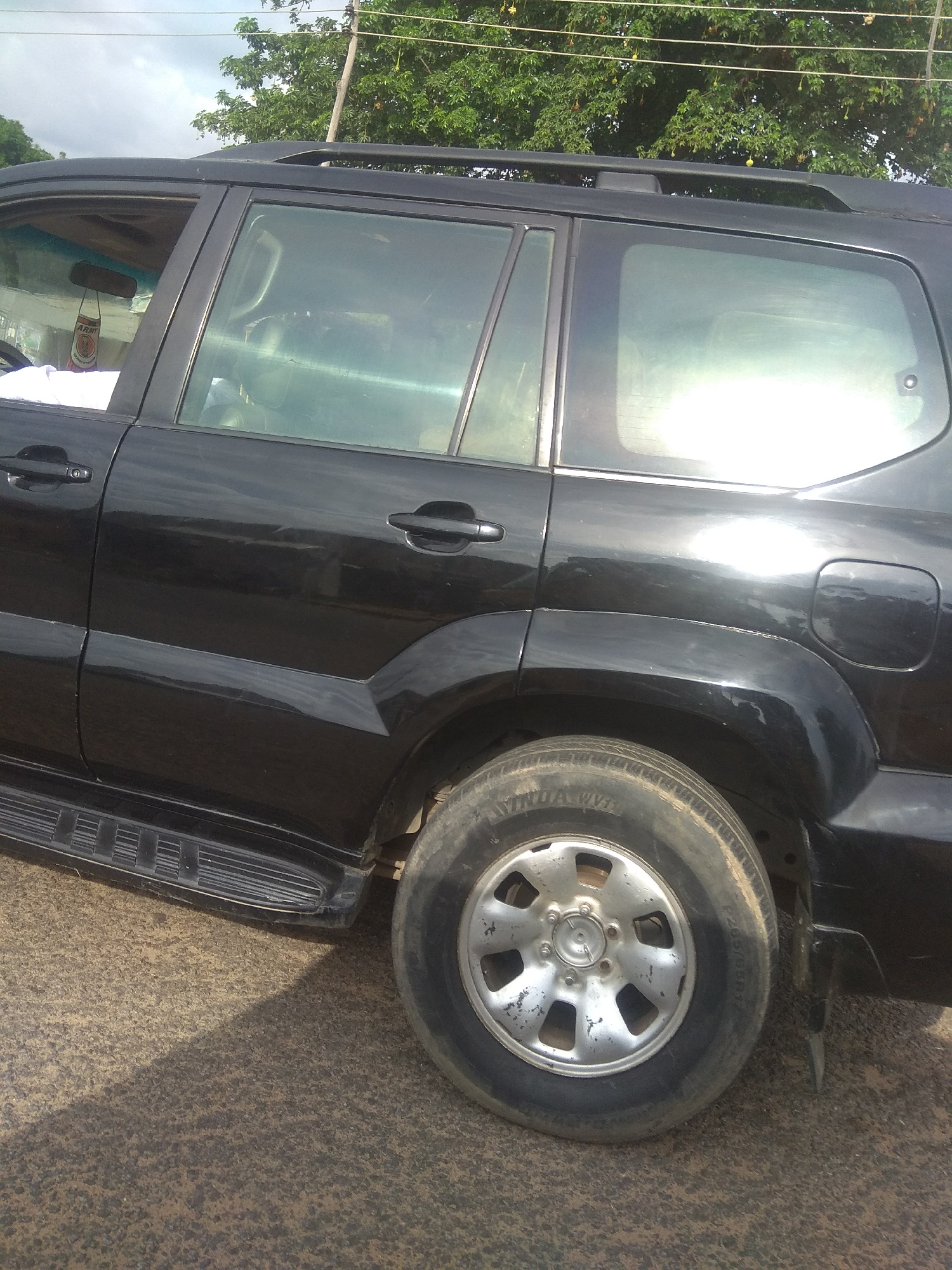 A black Toyota Land Cruiser Prado is parked on a paved road. The car is viewed from the side, with the rear passenger door and wheel prominently visible. The car's windows are tinted, reflecting the sky and surrounding trees. A red and white air freshener hangs from the rearview mirror inside the car. The environment appears to be an outdoor setting, possibly a roadside in Jalingo, Nigeria, with trees and power lines visible in the background. The road surface is rough and textured.