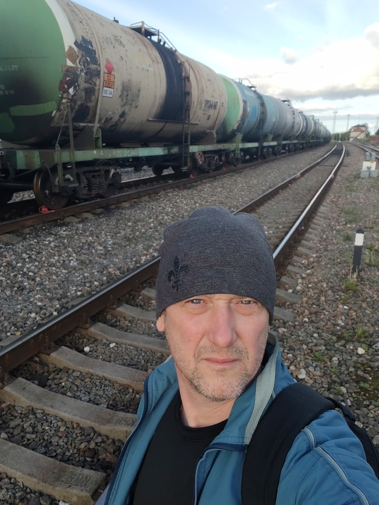 The image shows a man standing near a railway track in Bol'shie Mkhi, Russia. The man is wearing a gray beanie with a fleur-de-lis symbol, a blue jacket, and a black backpack. Behind him is a train composed of tank cars. The train is stopped on the tracks, which are surrounded by gravel. A white and black marker post stands near the track in the background. The sky is cloudy.
