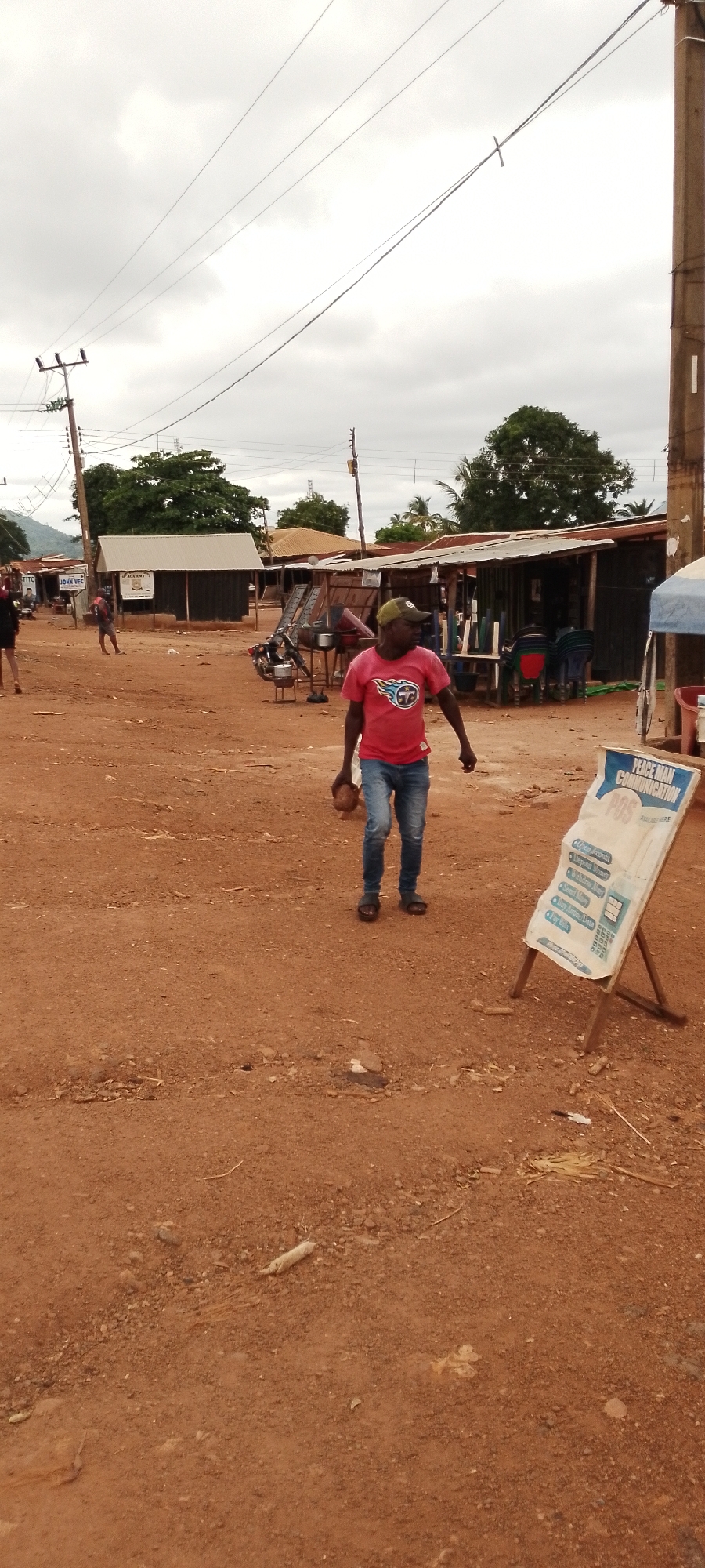 The image depicts an outdoor scene in a dusty, possibly rural or semi-urban environment, consistent with locations in Gboko, Nigeria. A man wearing a red t-shirt with a logo and blue jeans walks towards the right of the frame, carrying what appears to be a coconut in his left hand. He is wearing a green cap. In the background, there are several open-fronted stalls or shops with corrugated metal roofs. Some chairs are visible stacked behind one of the stalls. Electrical poles and overhead wires are present, indicating the availability of electricity. A sign on a wooden easel stands in the foreground on the right, bearing text related to "PEACE MAN COMMUNICATION" and listing services like "Open Product," "Internet Money," "Send Money," "Buy Airtime/Data," and "Pay Bills." Other individuals are visible in the background, appearing to be walking or standing near the stalls. The ground is covered in reddish-brown dirt with scattered debris. The sky is overcast, suggesting cloudy weather.