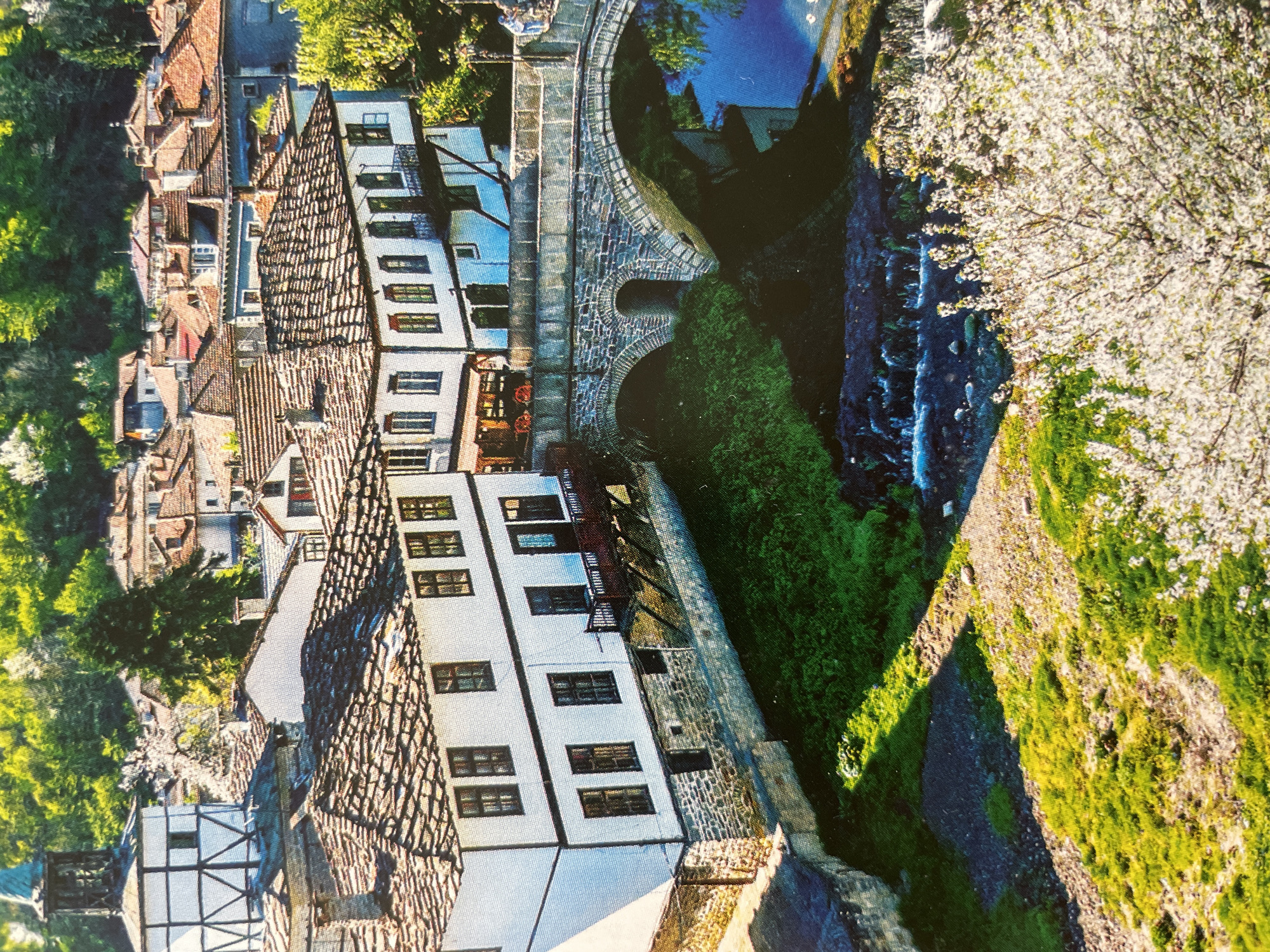 This image presents a high-angle view of a picturesque, historic town built along a winding river, characteristic of many settlements in Bulgaria. The scene is vibrant with natural light, suggesting a clear, sunny day.
In the upper and left portions of the frame, numerous traditional houses are clustered together on a steep hillside. These buildings feature distinctive red-tiled roofs, often gabled, and light-colored facades, many of which are white. Dark-framed windows are visible on most structures, and some show architectural details like balconies and timber framing, typical of Bulgarian Revival period architecture.
Centrally located, a beautifully preserved stone arch bridge spans the river. Its robust construction and arched design are prominent, connecting the settled area with the opposite bank. The river itself flows through a deep, verdant gorge. The water appears clear, with ripples and reflections, indicating its movement.
The right and lower sections of the image are dominated by lush greenery. The river banks are covered with dense green foliage, including grass and various trees. A particularly striking feature is a large tree in full white bloom, adding a touch of spring or early summer beauty to the landscape. Sunlight casts distinct shadows across the buildings and terrain, highlighting the textures of the tiled roofs and the stone bridge.
No people or discernible text are visible in the image. The overall atmosphere is one of tranquility and historical charm, showcasing a well-preserved architectural and natural landscape that aligns with the scenic beauty found in areas like Sofia's wider region or other historic Bulgarian towns.
