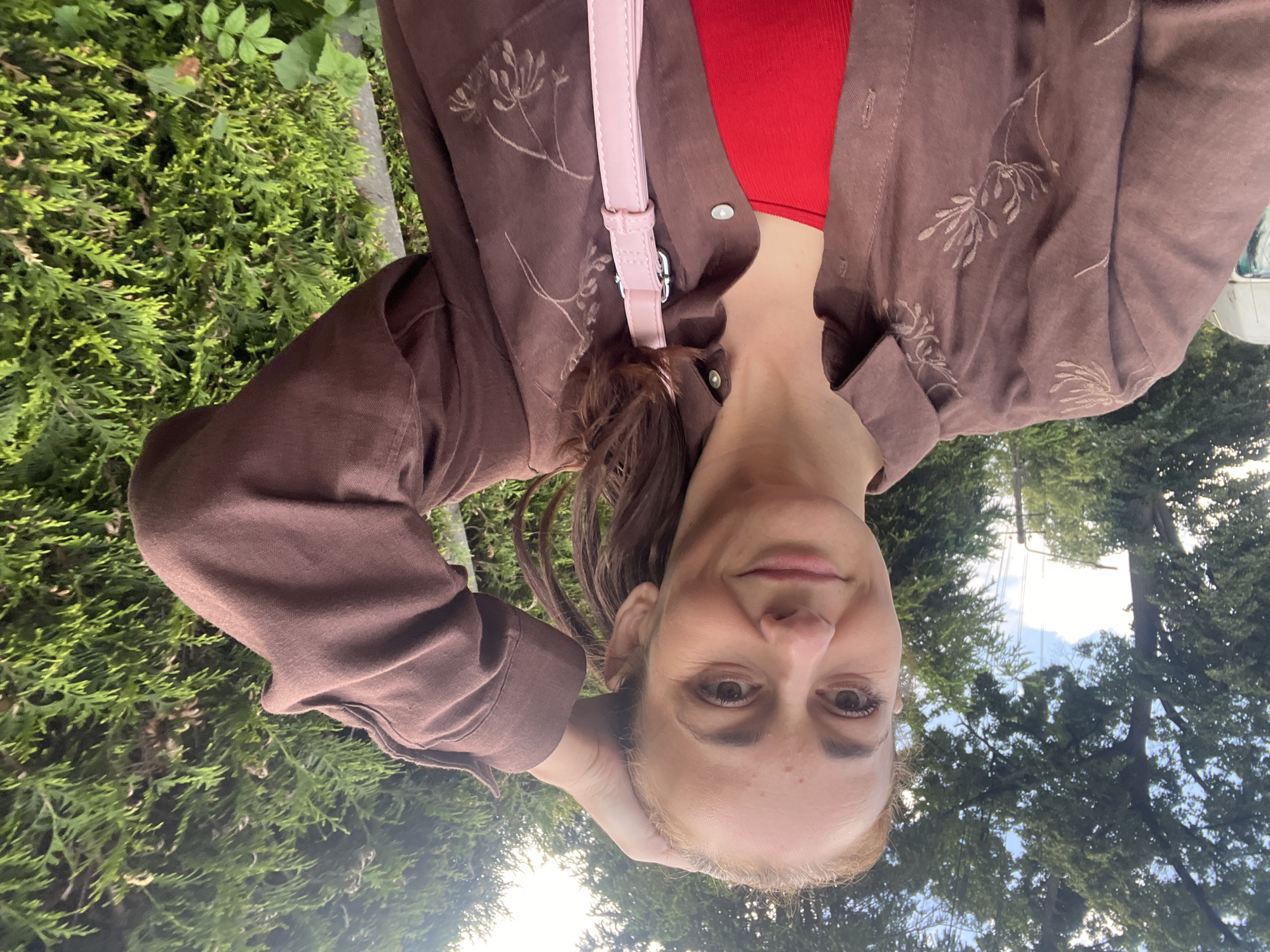 In an outdoor, daytime setting, a woman with light brown hair tied back in a ponytail is captured in a selfie. She is wearing a brown button-down shirt with embroidered floral designs and a red tank top underneath. A light pink crossbody bag with a silver buckle is visible. The woman is holding her right hand to her head, and her left arm is bent. She is looking at the camera with a slight smile. The background consists of lush green trees and foliage, suggesting a natural or park-like environment.