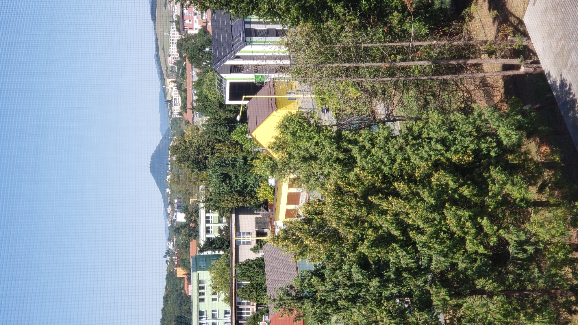 The image displays a cityscape with a prominent conical hill in the background, identified as the Kapušiansky hrad hill near Prešov, Slovakia. In the foreground, a cluster of evergreen trees partially obscures the view of several buildings. Among these structures is a bright yellow house with a brown roof and white window frames, and a modern building with a striped facade featuring green and white vertical stripes, solar panels on its roof, and a tall, dark rectangular structure at its side. Adjacent to the yellow house, a building with a brown tiled roof and visible white garage doors is situated. Further back, several larger, multi-story buildings are visible, including a green facade building with numerous windows. The sky is clear and blue. The overall scene suggests a suburban or semi-urban residential area with a mix of older and modern architecture, set against a natural hilly landscape.