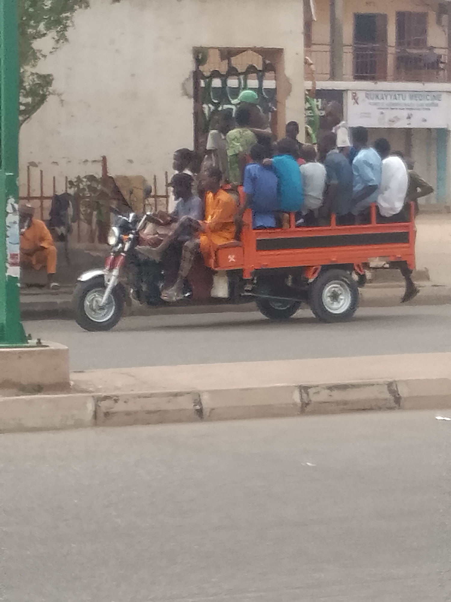 The image depicts an orange three-wheeled vehicle, possibly a modified motorcycle or auto-rickshaw, overloaded with numerous people. It is traveling on a paved road in an urban area. A person sits in a chair and is sitting next to a light pole on the left side of the road. In the background, there is a building with a sign visible that reads "RUKAYYATU MEDICINE". The general setting appears to be a busy, developing-world street. Based on the prompt, this is located in Pantame, Nigeria.