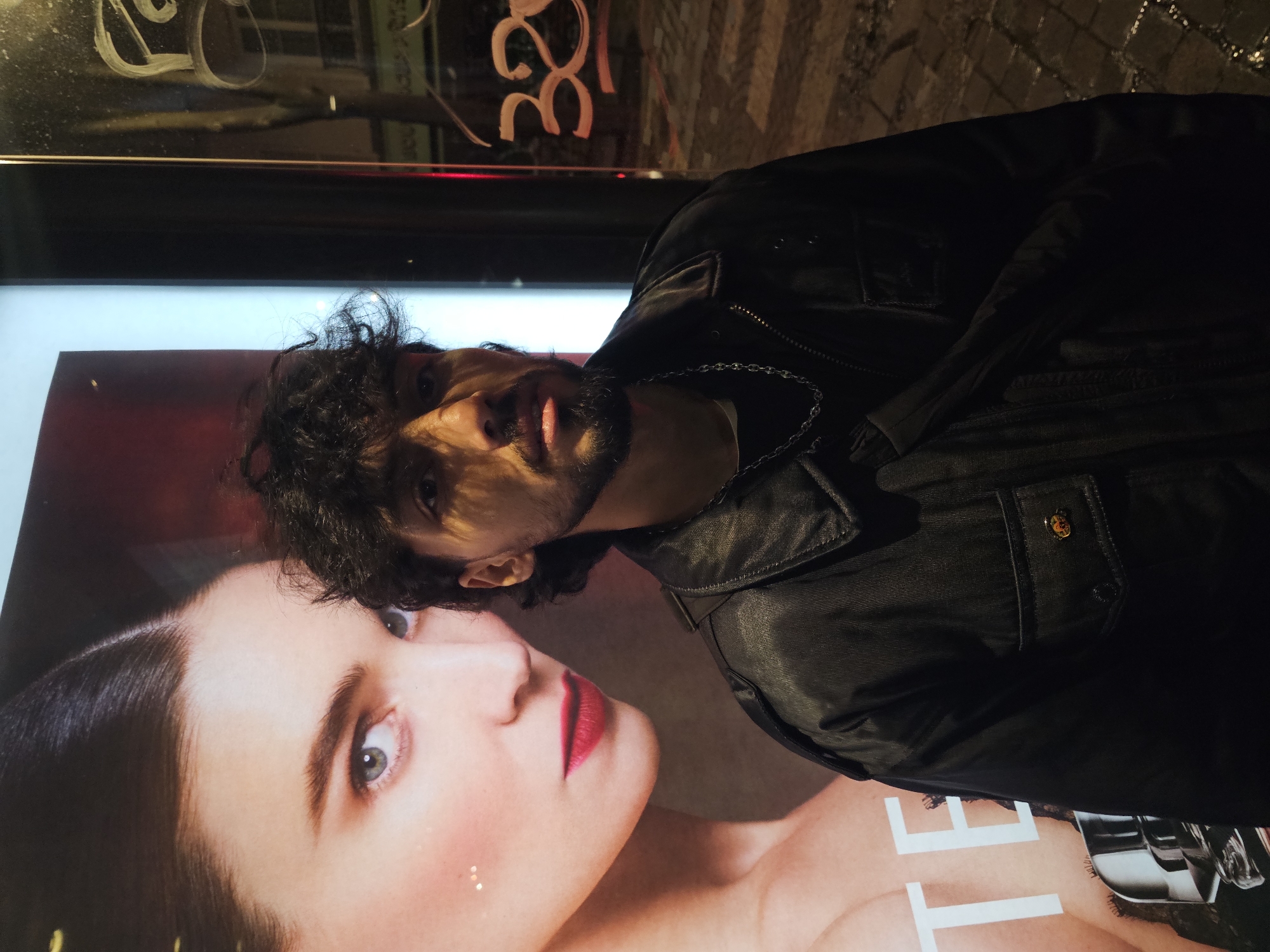 A medium shot shows a man standing in front of an illuminated advertisement, likely a poster displayed in a glass-fronted bus shelter or shop window. The man is of South Asian descent, with dark curly hair and a trimmed beard. He is wearing a black leather jacket and a silver chain is visible around his neck. He is looking directly at the camera with a neutral expression.
The advertisement features a close-up of a woman with fair skin and red lipstick, who appears to be looking away from the viewer.
The location is potentially in Paris, France. Reflected in the glass above the poster is the number "32," and a partially visible word that could be the name of a street. The background shows a cobblestone-paved area, typical of Parisian streets. The overall lighting suggests it is nighttime.