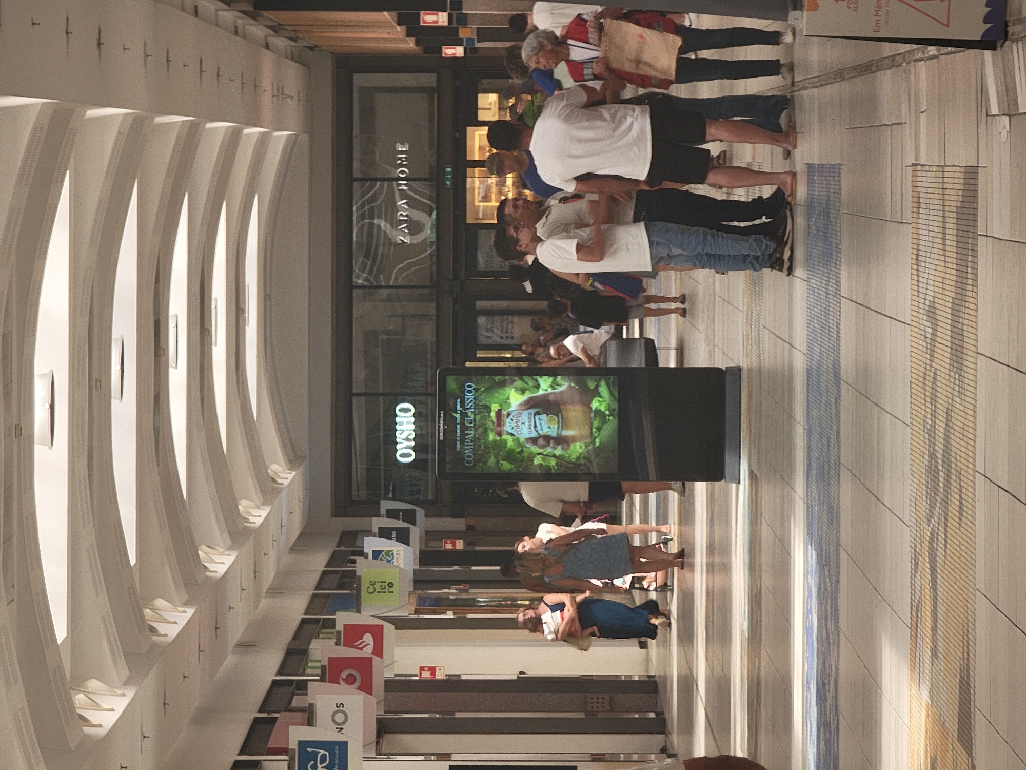 The image depicts the interior of a shopping mall, likely in Faro, Portugal, given the context of other images from the same source. The architecture features a high, arched ceiling with recessed lighting. Several retail storefronts are visible, including a Zara Home store with its logo prominently displayed. In the foreground, a digital advertising kiosk displays an advertisement for "COMPAL CLASSICO" juice. A group of people are gathered in the middle ground, some standing and conversing, while others are walking. One person in the foreground is walking away from the camera, with a stroller visible behind them. The floor is tiled with a patterned design. The overall scene conveys a typical day at a shopping center with shoppers browsing and interacting.