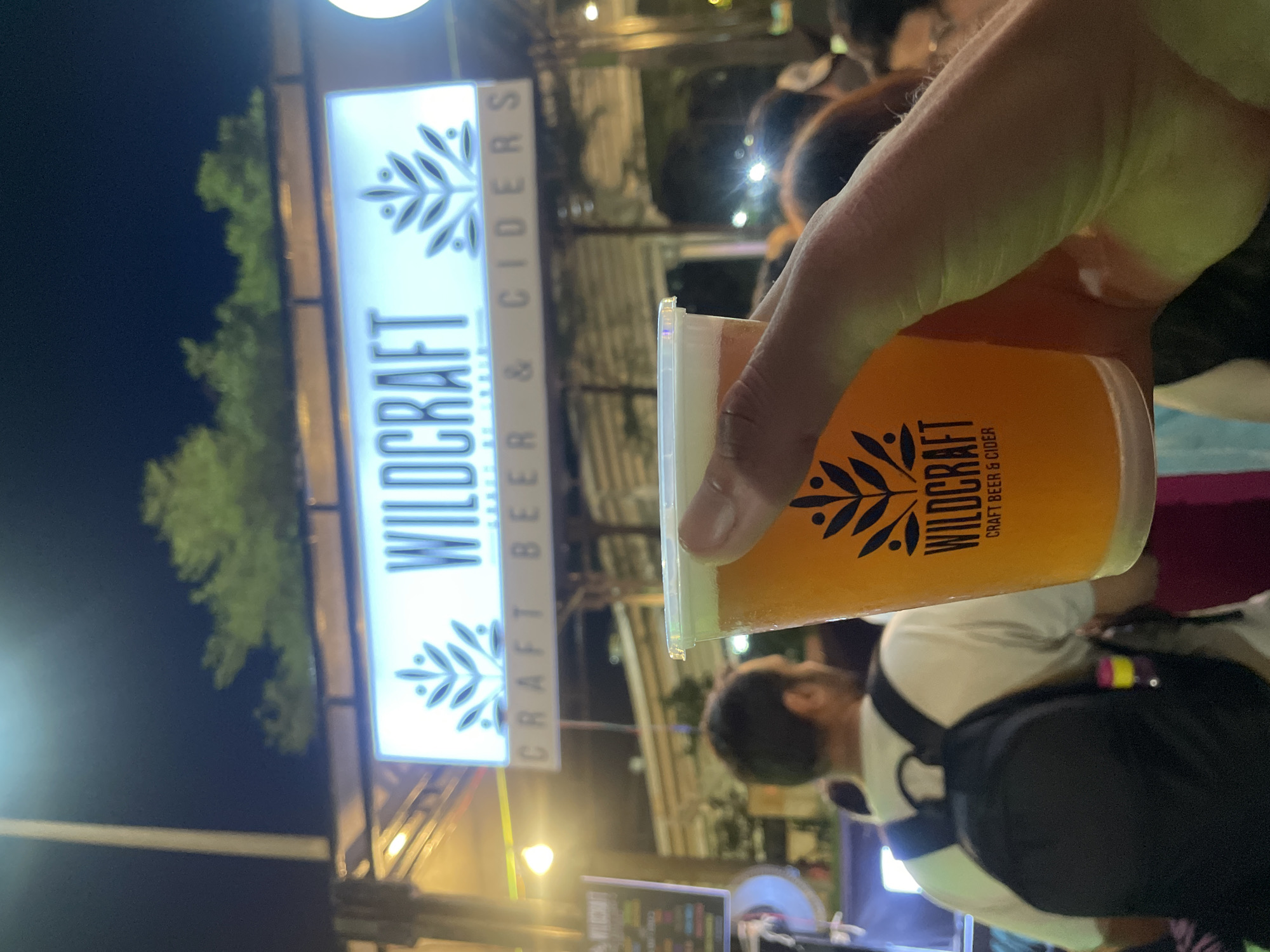 Here's a detailed description of the image:
**General Overview:** The image depicts a person holding a glass of beer or cider in front of a stall or venue named "Wildcraft." The scene appears to be taken at an outdoor event or market at night.
**People:**
* One person's hand is clearly visible, holding the glass.
* Other individuals are blurred in the background, suggesting a crowd. At least one person is wearing a white shirt and a backpack.
**Subjects/Objects:**
* **Drink:** The glass is filled with a yellow/orange-colored beverage, presumably beer or cider. The glass has the "Wildcraft" logo on it.
* **Signage:** A lit-up sign for "Wildcraft Craft Beer & Ciders" is prominent in the background.
* **Venue:** A stall or tent-like structure is implied by the lighting and the presence of other stalls (partially visible in the background).
* **Other Objects:** There are indistinct shapes and lights that suggest other stalls and equipment.
**Actions/Interactions:** The primary action is the holding of the drink. It is implied that the person has just purchased the drink or is about to consume it.
**Environment:** The setting is outdoors at night. There are trees visible.
**Location:** I can not determine the specific location, as there is no supporting evidence.