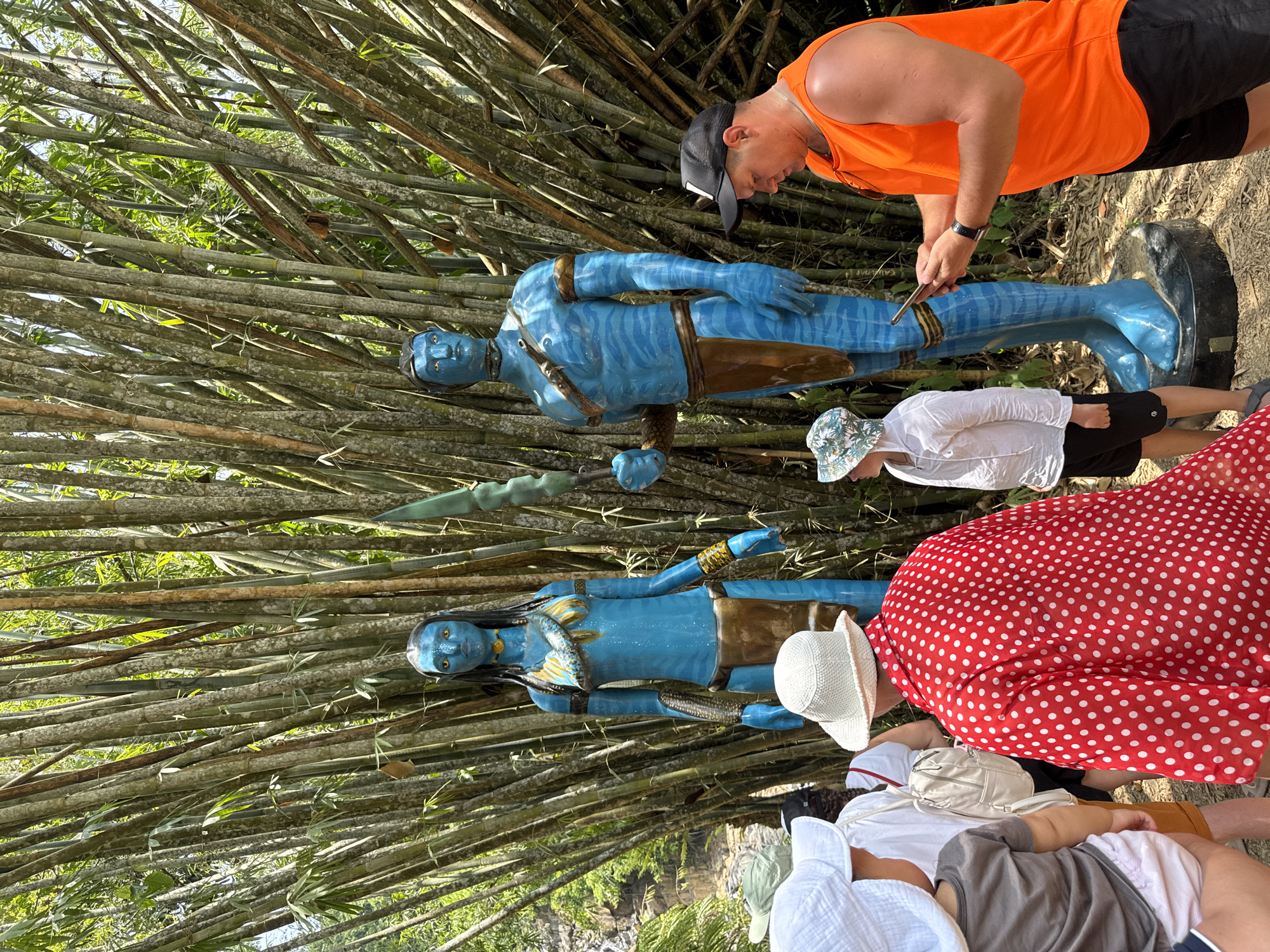 The image shows a group of people and two Avatar-like statues standing outdoors. The statues, painted in blue, are positioned in front of a dense thicket of bamboo. A man wearing a bright orange tank top and a black baseball cap is interacting with the upper statue. To his left, a child in a white shirt and patterned hat stands near the statue. Adjacent to them, a person in a red polka-dot dress and others wearing hats are also present. The location appears to be Ban Khlong Khian, Thailand, based on the presence of bamboo and the overall vegetation.