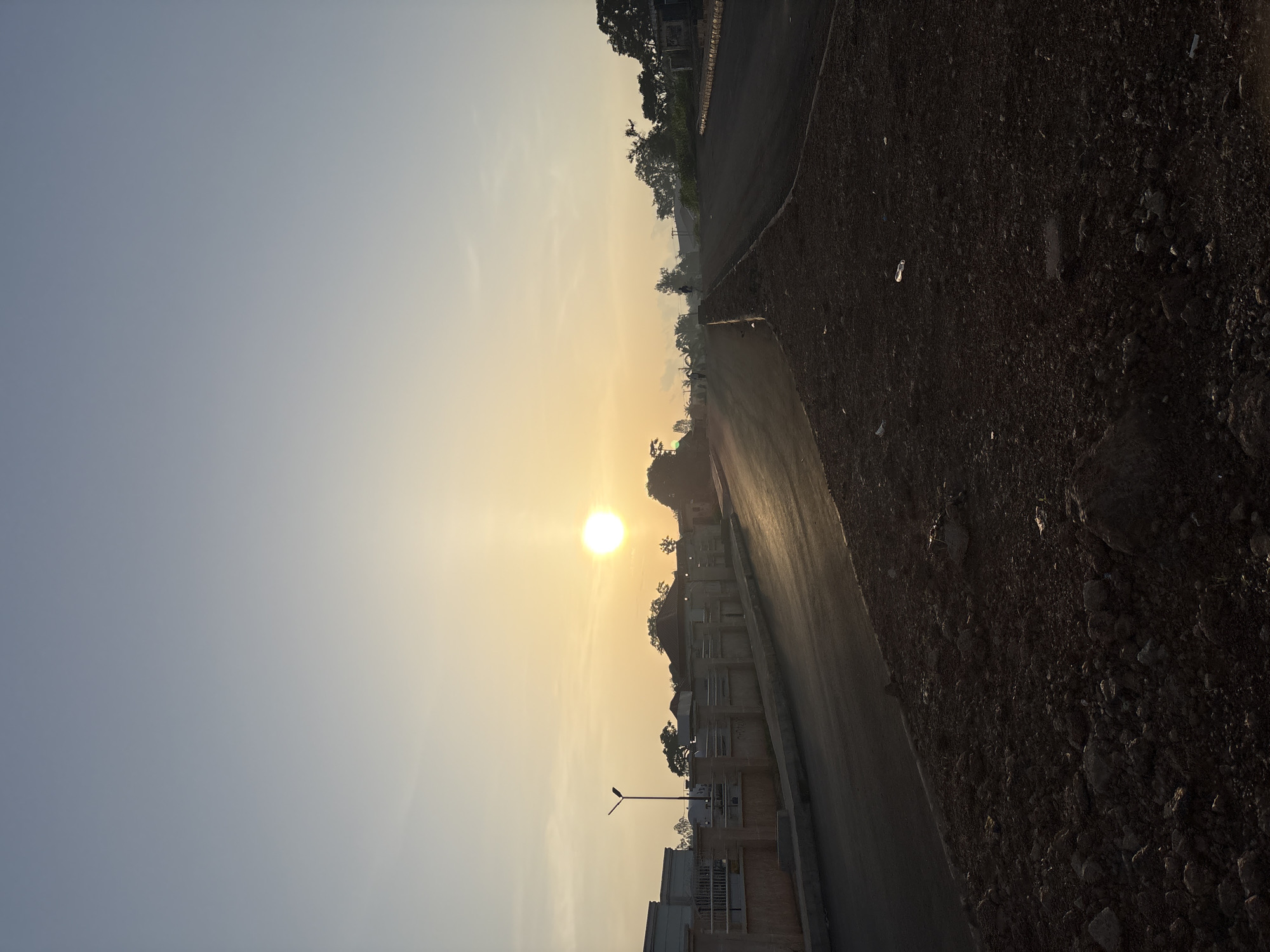 The image depicts a street scene at sunrise in Bukuru, Nigeria. The sun is low in the sky, casting a warm glow over the landscape. A paved road curves through the scene, with a dirt embankment on the right side and a paved sidewalk with a fence and buildings on the left. Trees and some structures are visible in the background. The sky is clear and light blue, with a gradient towards yellow near the horizon. There are no visible people in the image. The overall impression is one of a developing urban area at the start of a new day.
