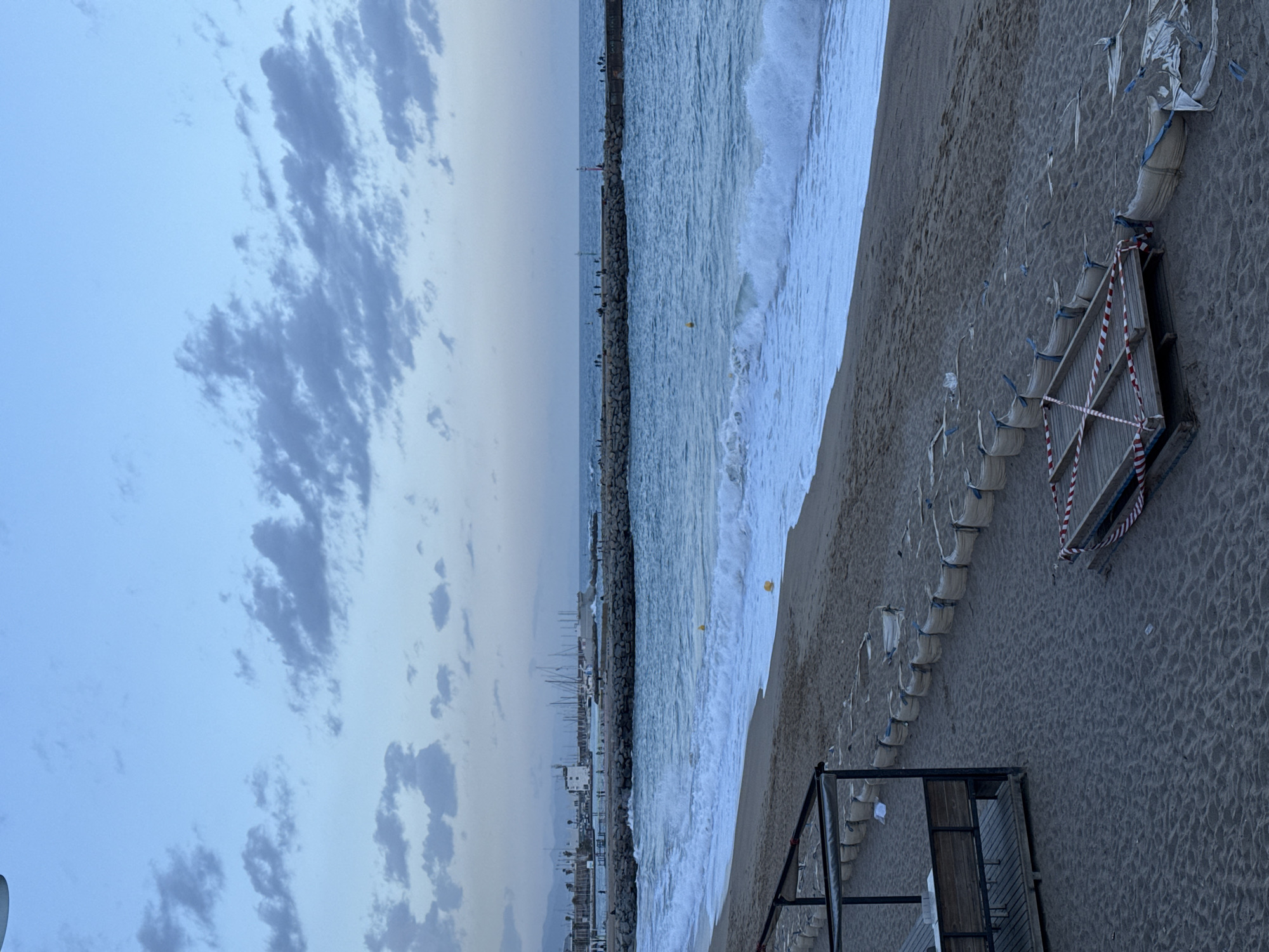 The image shows a beach scene in Barcelona, Spain, likely taken during the early morning or evening due to the subdued lighting. The beach slopes downwards from the right side, towards the water, where there are waves crashing onto the shore. A stone breakwater extends into the sea in the background, with possible harbor structures visible along it. The sky is overcast with gray and white clouds. In the foreground on the beach, there are rows of empty sandbags and construction materials, indicating beach reinforcement or repairs. There is no human activity visible.