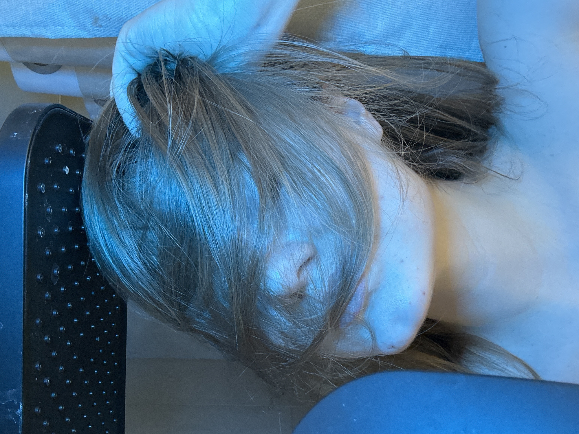 A close-up image depicts an individual under a black, rectangular shower head featuring multiple silver-colored nozzles, located in a bathroom in Kazan, Russia. The person's long, light brown hair falls across their face, obscuring the eyes and forehead, but revealing the nose, upper lip, chin, and part of the neck and shoulders. The individual's left hand, visible on the right side of the frame, is raised and grasping a section of hair near the top of the head. To the right, a light-colored, textured material, possibly a shower curtain or tiled wall, is partially visible alongside a vertical support structure. A blurred blue object is present on the far left of the frame. The entire scene is illuminated by a dominant blue light source.