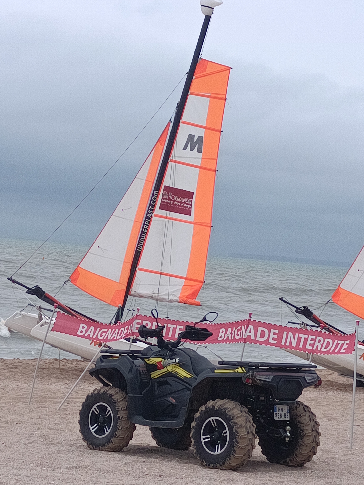 The image shows a beach scene in Houlgate, France, on an overcast day. In the foreground, a black ATV is parked on the sand. Behind the ATV, two small sailboats with orange and white sails are positioned on the beach. A pink banner with white text reading "BAIGNADE INTERDITE" (Bathing Forbidden) is stretched between two poles, obscuring part of the sailboats. The sea is visible in the background, with gentle waves. The sky is cloudy, suggesting potentially inclement weather.