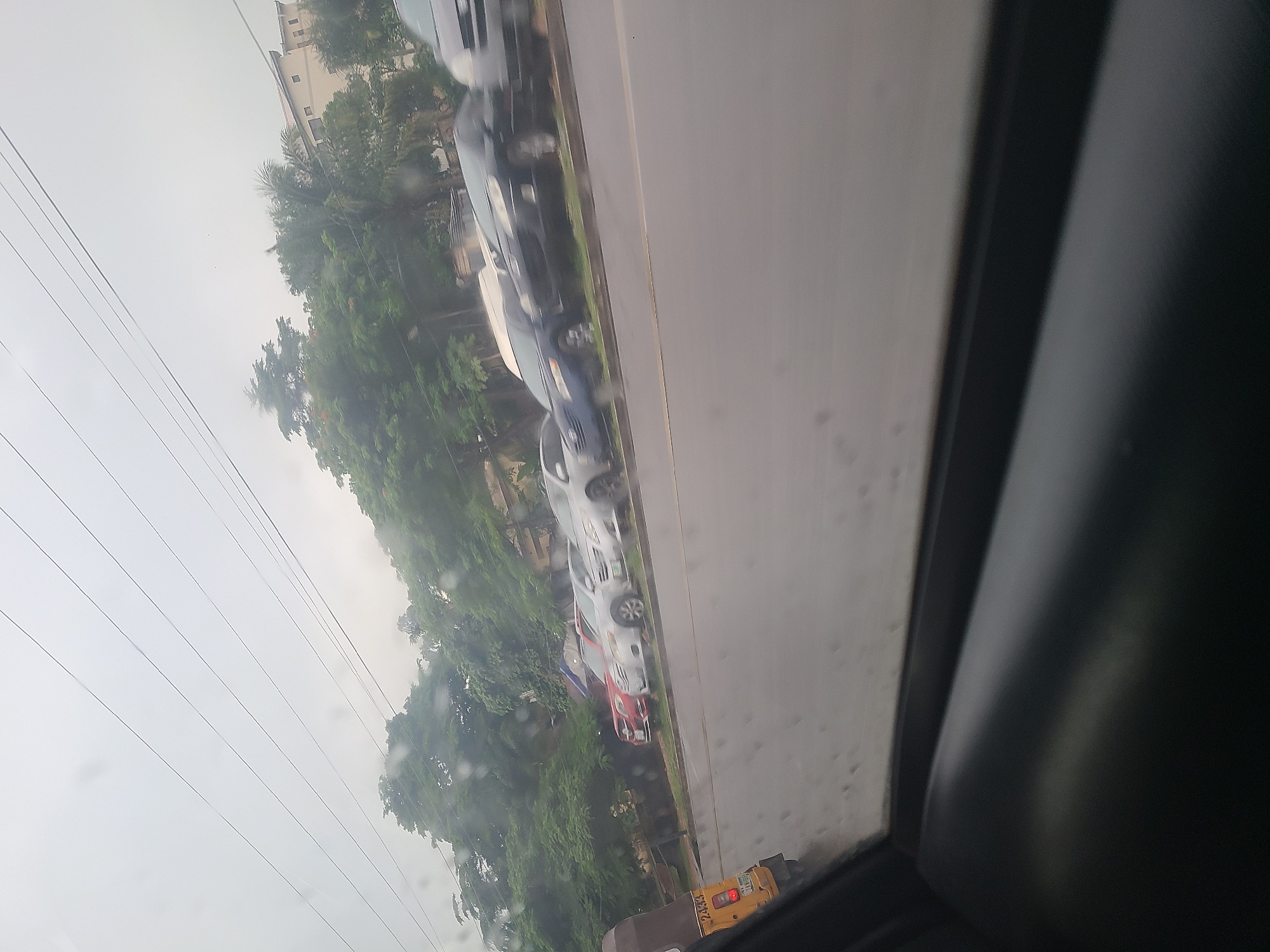The image shows a street scene in Karu, Nigeria, viewed from a car window. Several cars are parked in a line along the side of a road. The weather appears to be overcast or rainy, as indicated by water droplets on the window. Lush green trees and some buildings are visible in the background. The overall atmosphere is one of a typical day in a busy urban or suburban area.
There are no people clearly visible in the image. The primary objects are the cars and the surrounding environment. The scene depicts a street with parked vehicles, suggestive of everyday traffic or a residential area. The presence of power lines in the sky further reinforces the urban setting.
Notable details include the overcast sky, suggesting it might be daytime, but with limited sunlight. The water on the window could indicate recent or ongoing rain. The trees suggest a tropical or subtropical climate. The presence of multiple cars lined up could be due to parking or a traffic jam, though the cars appear stationary.
A small yellow vehicle with the license plate "LZ4844" is visible towards the bottom right of the image. No other discernible text is present. The angle of the photo, being taken from inside a vehicle and tilted, gives a dynamic perspective to the scene.
