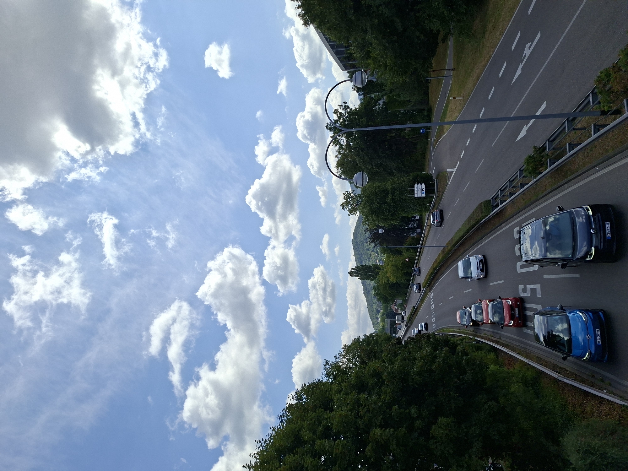 This media file captures a daytime scene on a highway in Baden-Baden, Germany, likely during fair weather given the presence of fluffy white clouds against a bright blue sky. The image is oriented vertically, showing a curved road with multiple lanes of traffic.
The main subjects are vehicles driving on the highway. These include a blue Volkswagen Golf, a black Volkswagen, several red cars, a white car, and other vehicles further in the distance. The road is marked with white lane lines and directional arrows, indicating a typical highway setup. Along the side of the road, there are streetlights with circular lamps and a metal guardrail separating the highway from the surrounding greenery. Dense trees line the side of the road, and a hillside covered in trees and some buildings is visible in the background.
The activity depicted is a flow of traffic on a highway. There are no people clearly visible in the frame. The time of day appears to be daytime, possibly late morning or afternoon, judging by the brightness of the sky and the shadows. The weather seems pleasant, with scattered clouds.
Visible text includes license plates on some of the cars. For example, the blue car has a license plate reading "B AWH 393E". The black car appears to have a plate starting with "MA". There are also road signs, some of which are partially visible and indicate traffic regulations or destinations, but the text is too small to read clearly. The overall impression is one of a busy but orderly highway in a scenic, possibly suburban or exurban, environment in Germany.
