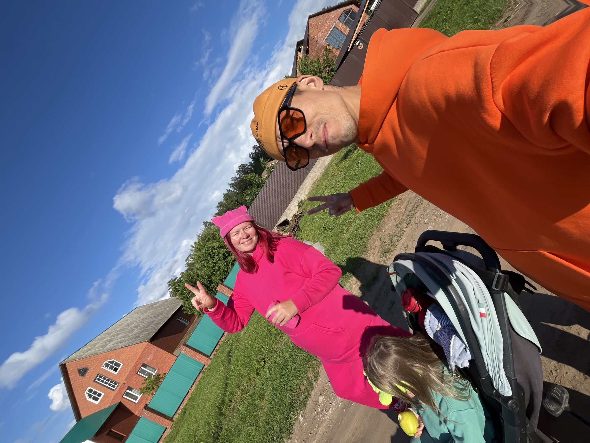 This tilted outdoor selfie captures three individuals enjoying a sunny day in what appears to be a rural or suburban setting in Chaikovsky, Russia. The bright blue sky with fluffy white clouds indicates clear, pleasant weather, likely midday or afternoon.
In the foreground on the right, a man wears an orange hooded sweatshirt, dark sunglasses, and a light brown beanie, which has some indistinct text. He looks towards the camera, making a peace sign with his right hand. To his left, a woman with vibrant pink hair, a matching pink beanie, and a pink full-body jumpsuit smiles widely, also flashing a peace sign. She holds a pink cup or bottle in her left hand.
Partially visible at the bottom right is a child with long, light-colored hair, wearing a light green top. The child is holding a yellow-green fruit, resembling an apple or pear, and is seated next to a black and light blue/grey baby stroller. The stroller features a small patch displaying the colors of the Italian flag.
The background reveals a dirt path, lush green grass, and residential buildings. A prominent two-story brick house with white-framed windows and green metal fences is visible on the left. Another house can be seen further back on the right. The cheerful expressions of the adults suggest a relaxed and happy family outing.