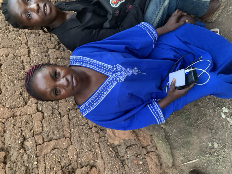 Two women sit outdoors. One wears a black shirt with a colorful emblem, along with jeans. The second, closer to the camera, dons a vibrant blue garment decorated with white embroidery around the neckline and sleeves, and holds a charging power bank with attached wires. They are positioned next to a cracked, earthen structure, likely a crumbling wall or path, with sparse vegetation surrounding them. The scene is captured under natural light.