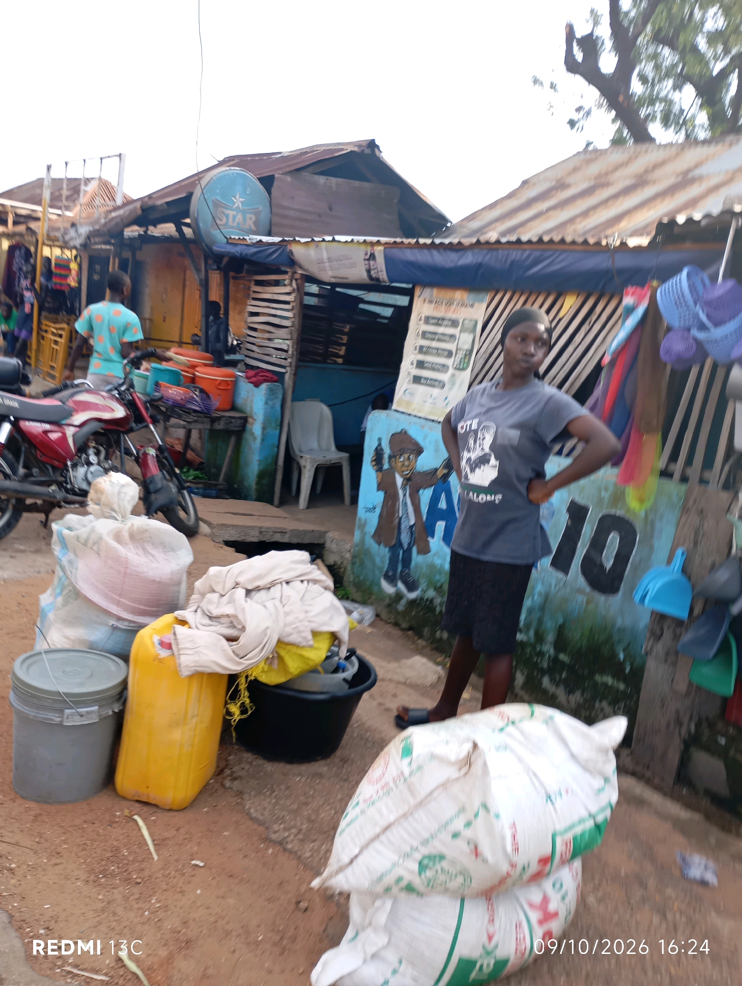 The image depicts an outdoor scene in Shendam, Nigeria, likely a marketplace or street-side vendor area. In the foreground, several large bags, one white with green lettering and another partially visible beneath it, are placed on the ground. To their left, a large grey bucket with a lid sits next to a yellow plastic jerrycan. A black plastic basin filled with items, including some fabric and a yellow bag, is positioned behind the jerrycan. Clothes and other assorted items are hanging on display in the background. A motorcycle is parked on the left side of the image. A young woman, wearing a grey t-shirt with a graphic and a black skirt, stands with her hands on her hips, looking to her left. Behind her, a building with a corrugated metal roof has various signs and graffiti on its wall, including a blue circular sign with "STAR" written in white letters. The ground is made of dirt or compacted earth. The lighting suggests it is late afternoon.