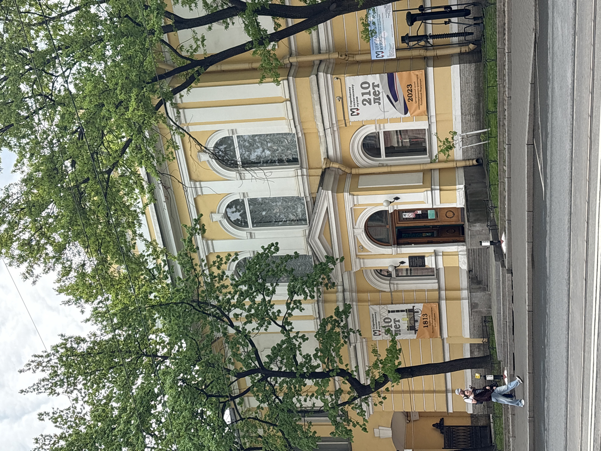 An outdoor, slightly high-angle shot captures a yellow and white neoclassical building, partially obscured by lush green trees, under an overcast daytime sky. The building features large arched windows with decorative white frames and columns, and a main entrance with a wooden door. A sidewalk and a street run parallel to the building.
Prominently displayed on the building's facade are several banners with text in Russian. One large banner reads "M ЦЕНТРАЛЬНЫЙ МУЗЕЙ ЖЕЛЕЗНОДОРОЖНОГО ТРАНСПОРТА РОССИЙСКОЙ ФЕДЕРАЦИИ" (Central Museum of Railway Transport of the Russian Federation). Other banners celebrate "M СТАРЕЙШЕМУ ТЕХНИЧЕСКОМУ МУЗЕЮ 210 ЛЕТ" (To the oldest technical museum, 210 years), with the years "1813" and "2023" also visible, indicating the museum's founding year and the current year of celebration. Industrial-looking equipment, possibly railway-related, is displayed on a ledge above one of the banners.
On the sidewalk near the street, a person wearing jeans and a dark jacket is sitting on a low wall or bench, looking towards the street. The street appears to be paved with asphalt, and there's a small patch of grass between the sidewalk and the street. The dense foliage of the trees fills the upper left portion of the frame, with sunlight filtering through the leaves. The scene depicts a quiet urban environment, likely in Saint Petersburg, Russia, given the architectural style and specific museum mentioned.