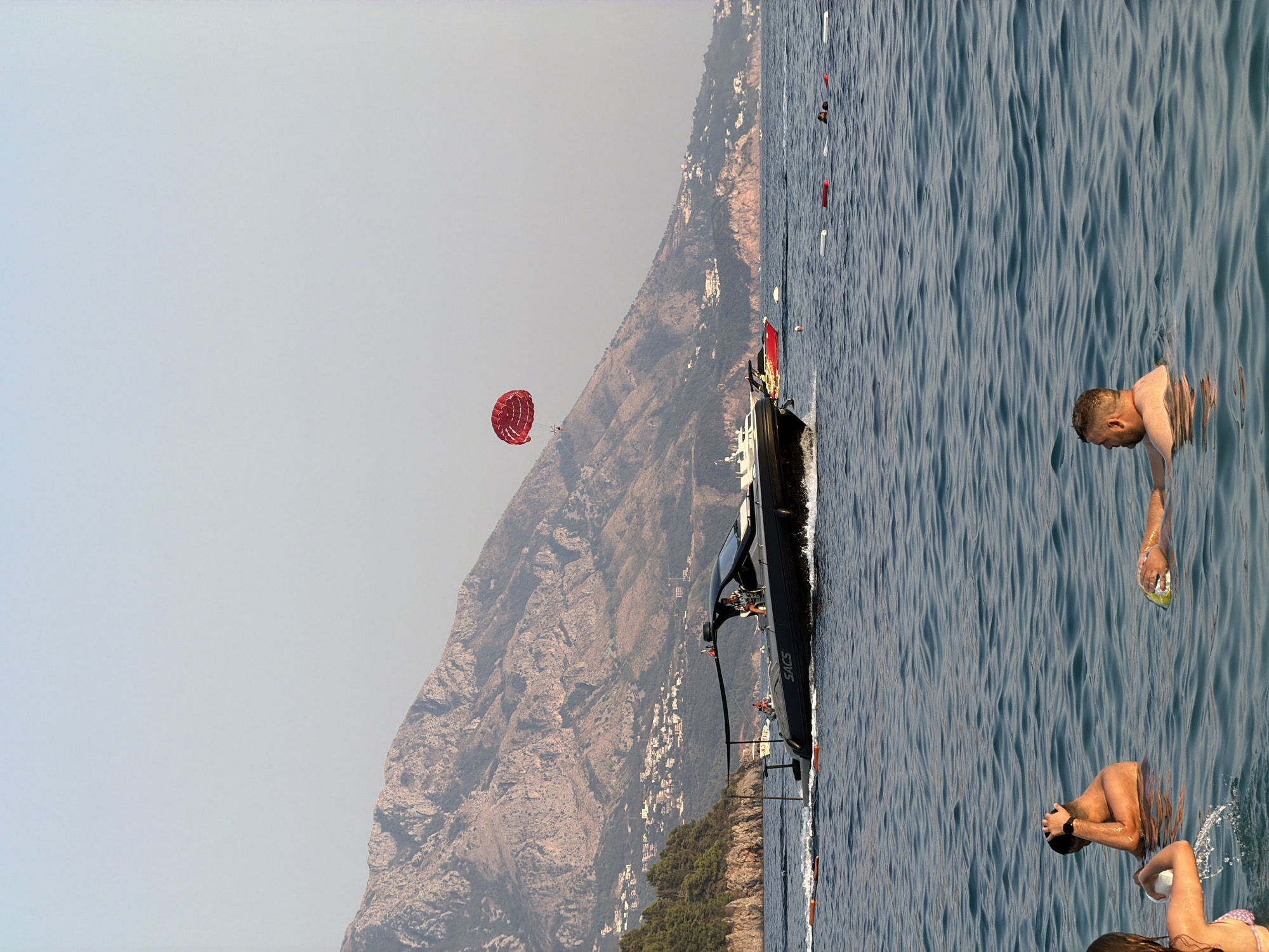 The image depicts a coastal scene likely near Budva, Montenegro, with a mountainous backdrop featuring scattered buildings. In the foreground, the choppy blue sea has several individuals swimming. One person is partially submerged, looking to the left, while another is floating on their back, holding a yellow object. A third person is visible in the lower left corner, facing away from the camera, with water splashing around them.
Further out in the water, a dark-colored speed boat with "SACS" branding is moving from left to right, creating a wake. A person is visible at the helm of the boat. In the distance, a red parachute is suspended in the air, with a person attached, suggesting parasailing activity. Another red boat is positioned behind the speed boat. The water is dotted with buoys. The sky is a hazy light blue, and the overall lighting suggests it is late afternoon.