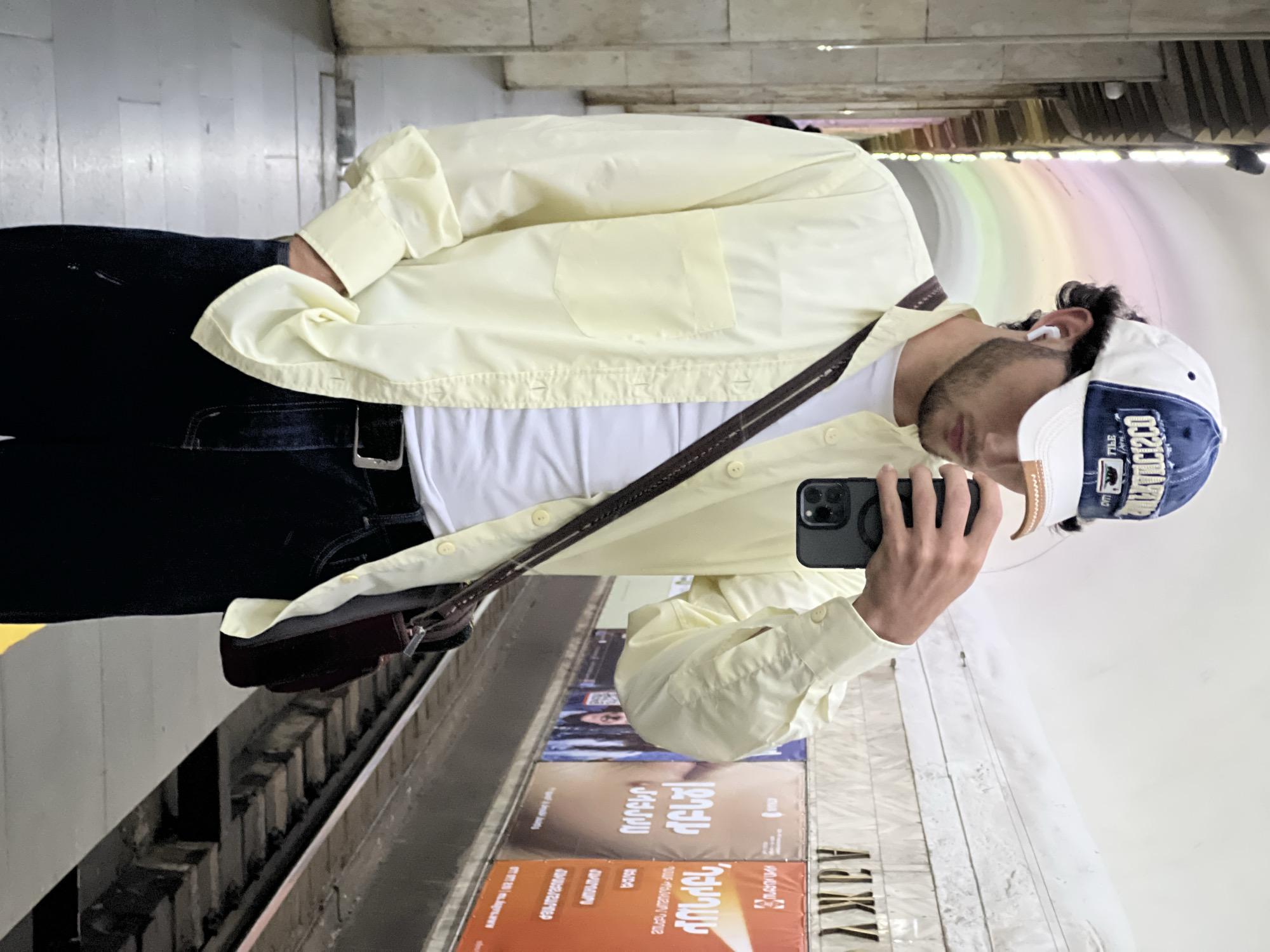 A person is taking a selfie in what appears to be a Yerevan subway station. The person is wearing dark jeans, a white t-shirt, and a light yellow button-down shirt. A brown bag strap crosses the person's torso. The person is also wearing a baseball cap. Subway advertisements can be seen on the wall behind the person. The ceiling of the subway tunnel has an overhead rainbow decoration.