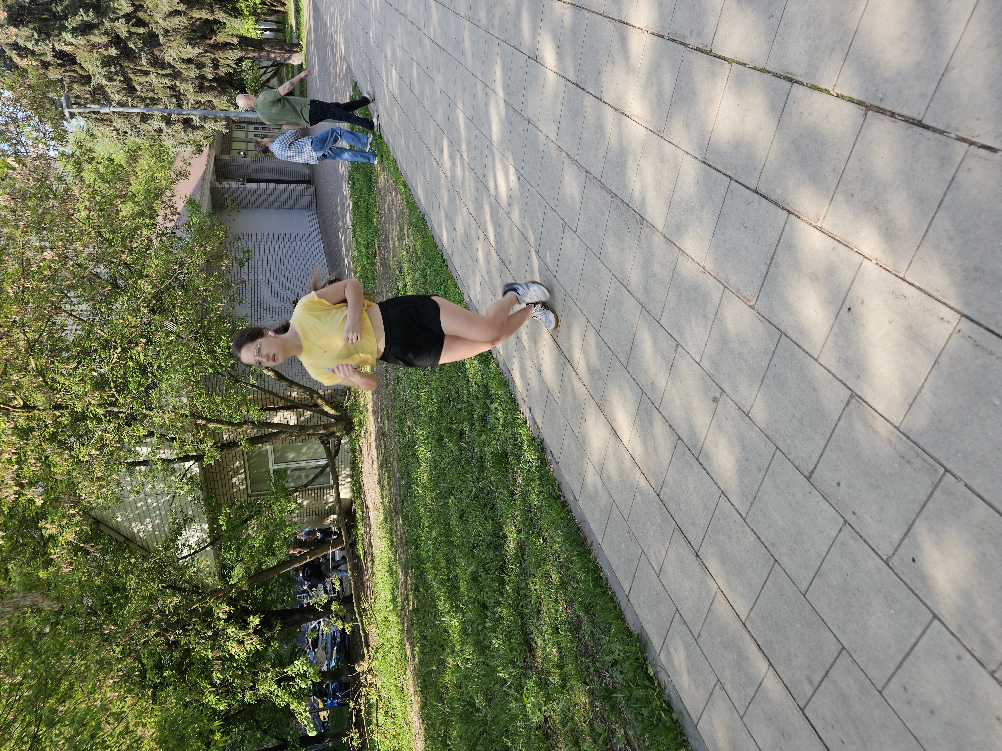The image shows a young woman standing on a paved walkway in Khimki, Russia. She is wearing a yellow t-shirt and black shorts and appears to be holding a cell phone. The walkway is lined with grass and trees. In the background, there are people standing near a light post in front of a building with gray brick. Cars are parked beside the trees. The lighting suggests it is daytime.