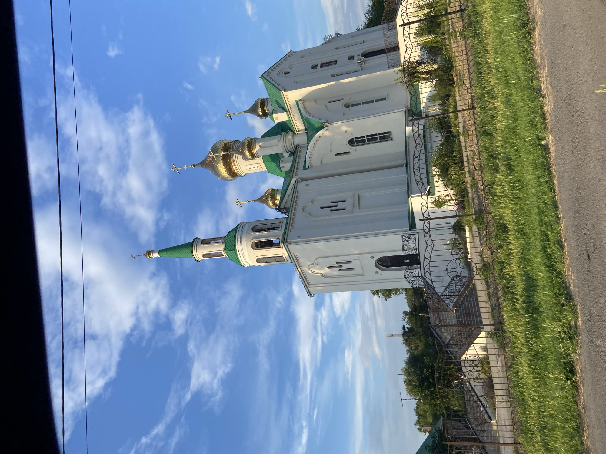 The image depicts an Orthodox church with white walls and green roofs, topped with golden domes. The church is situated in a rural or suburban setting, with a grassy area and a metal fence bordering the property. A paved road is visible to the right, and a telephone pole and wires are present in the foreground. The sky is a clear blue with scattered white clouds, suggesting a daytime scene, likely in the late afternoon or early evening due to the warm lighting. There are no people visible in the image. The overall impression is one of peacefulness and architectural beauty. The image is oriented vertically, with the church structure running from the bottom left to the top right of the frame.