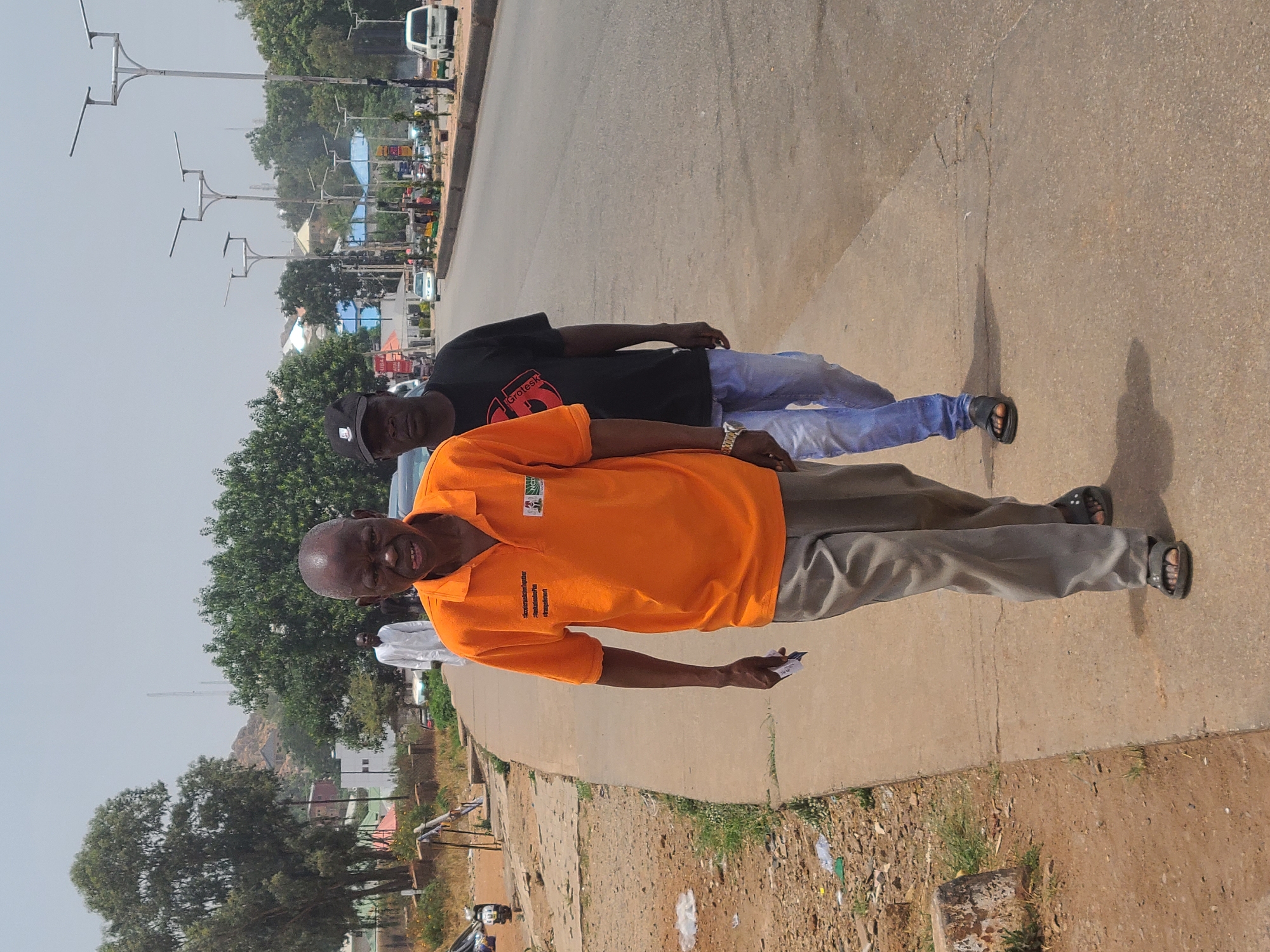 Two men are standing on the side of a road. The man in the foreground is wearing an orange shirt and gray pants, and holding a small, white object in his right hand. He is smiling. The man behind him is wearing a black shirt, blue jeans, and a black baseball cap. They are both wearing sandals. The road is paved, with a sidewalk on the left side. There are trees and buildings in the background. Streetlights are visible on the opposite side of the road.
Based on visual cues of the urban environment and knowledge of typical street scenes, this image is likely located in the city of Jos, Nigeria. Specifically, the style of streetlights and the roadside vegetation are characteristic of Jos.