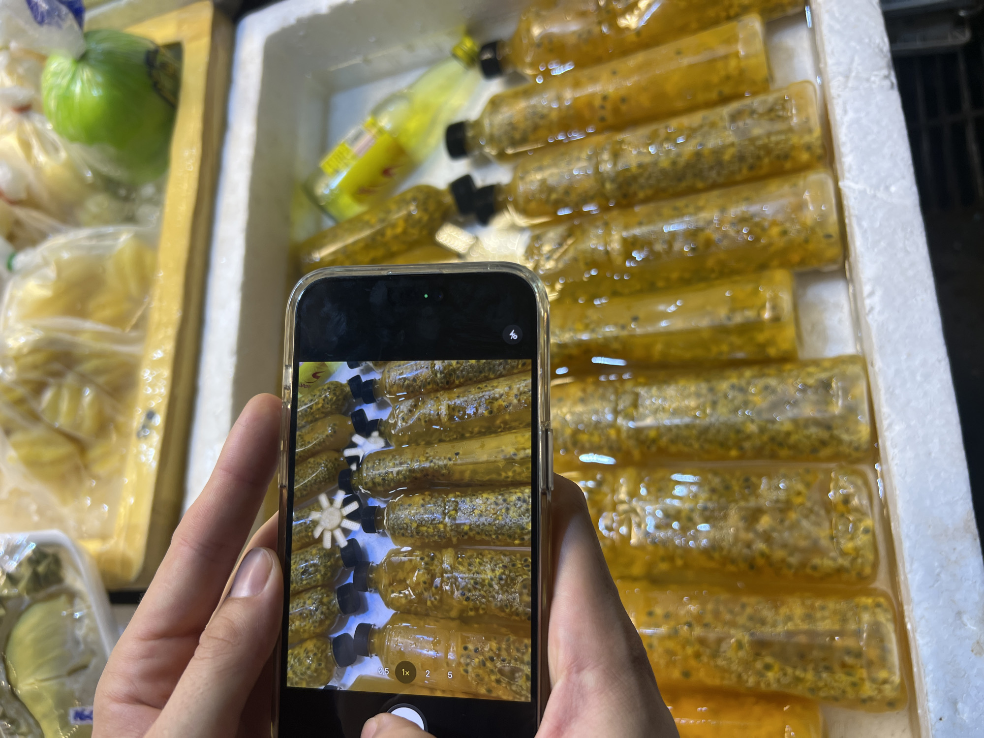 A person is holding a smartphone, taking a picture of bottles filled with passion fruit juice. The bottles are arranged in a white styrofoam container, likely at a market. The juice is a vibrant yellow with visible passion fruit seeds. In the background, there are other items, including a green fruit in a plastic bag and other bottles of what appears to be a yellow beverage. The scene suggests a bright, possibly daytime setting, typical of a local market in Thailand. There is no visible text in the image. The focus is on the product being photographed, highlighting its visual appeal.