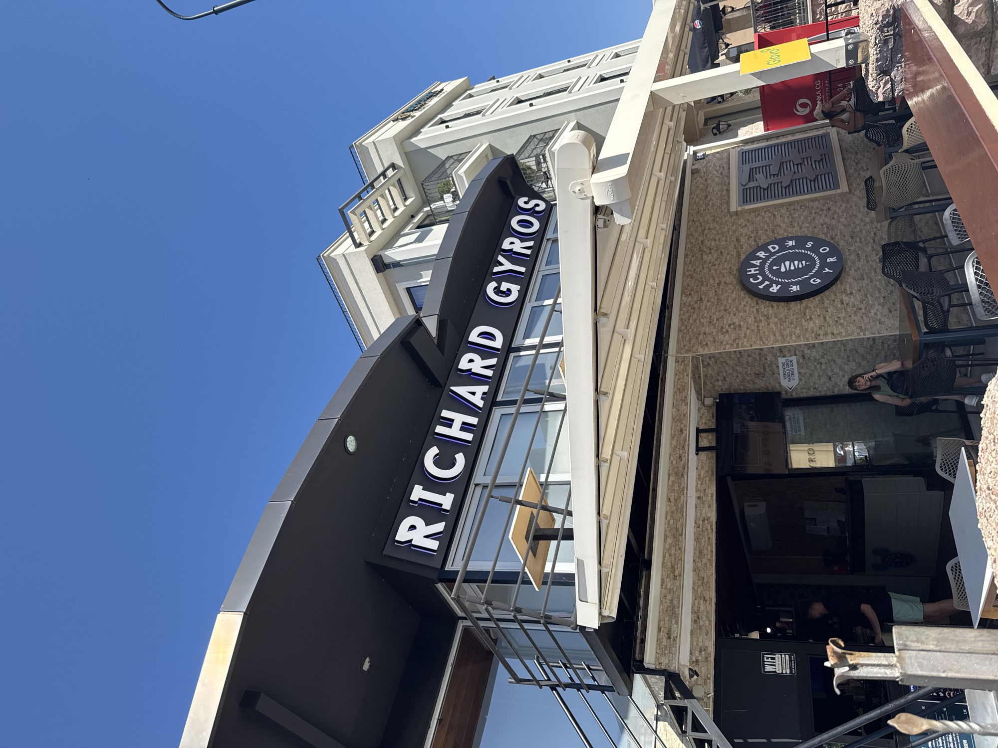 A low-angle, exterior medium shot captures a modern building in Budva, Montenegro. The building features a prominent black awning with illuminated white lettering that reads "RICHARD GYROS." Below this, a balcony area with tables and chairs is visible, protected by a metal railing. The facade of the building is a mix of glass, metal, and stone.
On the ground level, there is an entrance to the establishment. A man in a black t-shirt and light green shorts is standing just inside, appearing to be working. A woman in a green tank top is seated at a table outside, talking on her phone. Several other tables and chairs are arranged on the patio. A circular sign with "RICHARD GYROS" written around a depiction of a gyro is mounted on the stone wall.
In the background, a taller, lighter-colored building rises against a clear blue sky. The overall scene suggests a bright, sunny day.