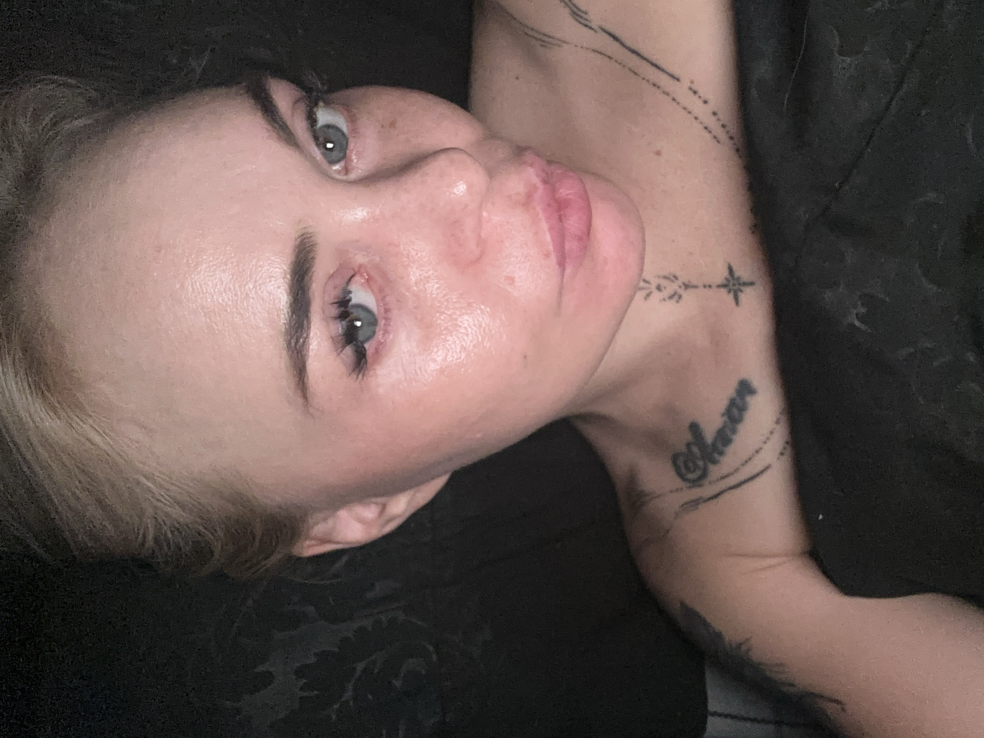 A close-up, high-angle, indoor, medium shot captures a woman with blonde hair pulled back from her face, as she lies in bed. The woman has light blue eyes, light skin with freckles, and appears to be wearing light makeup. She has several tattoos visible on her upper body, including on her left arm, collarbone, and chest. The background is dark and out of focus, with some pattern visible. The woman is looking directly at the camera with her lips pursed slightly. The lighting is dim, and the overall tone of the image is intimate.