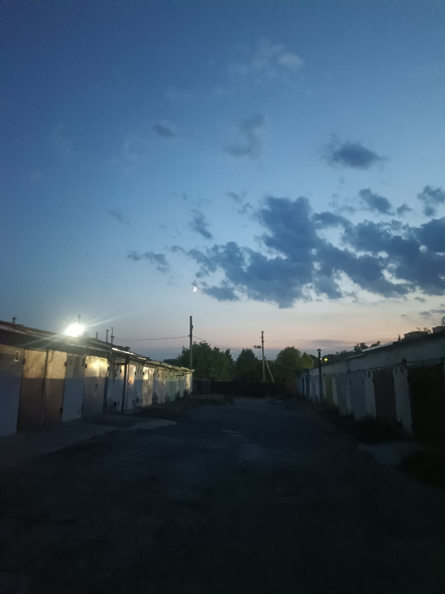 The image depicts an outdoor scene at dusk in a location that appears to be an alleyway or street lined with garages. The sky is a deep blue, transitioning to a lighter hue near the horizon, with scattered clouds. A crescent moon is visible in the upper portion of the sky. The garages are arranged in two rows, facing each other, with a dark, unpaved ground surface in between. The garage doors are varied in color and condition, with some showing signs of wear. Artificial lights illuminate some of the garages, casting bright flares. Trees and vegetation are visible in the background, suggesting an area with some greenery. A utility pole with visible wires is also present. The scene is devoid of people and any specific actions are not discernible. The overall atmosphere is quiet and somewhat desolate. Based on the provided information, the location is identified as Rybnitsa, Moldova.