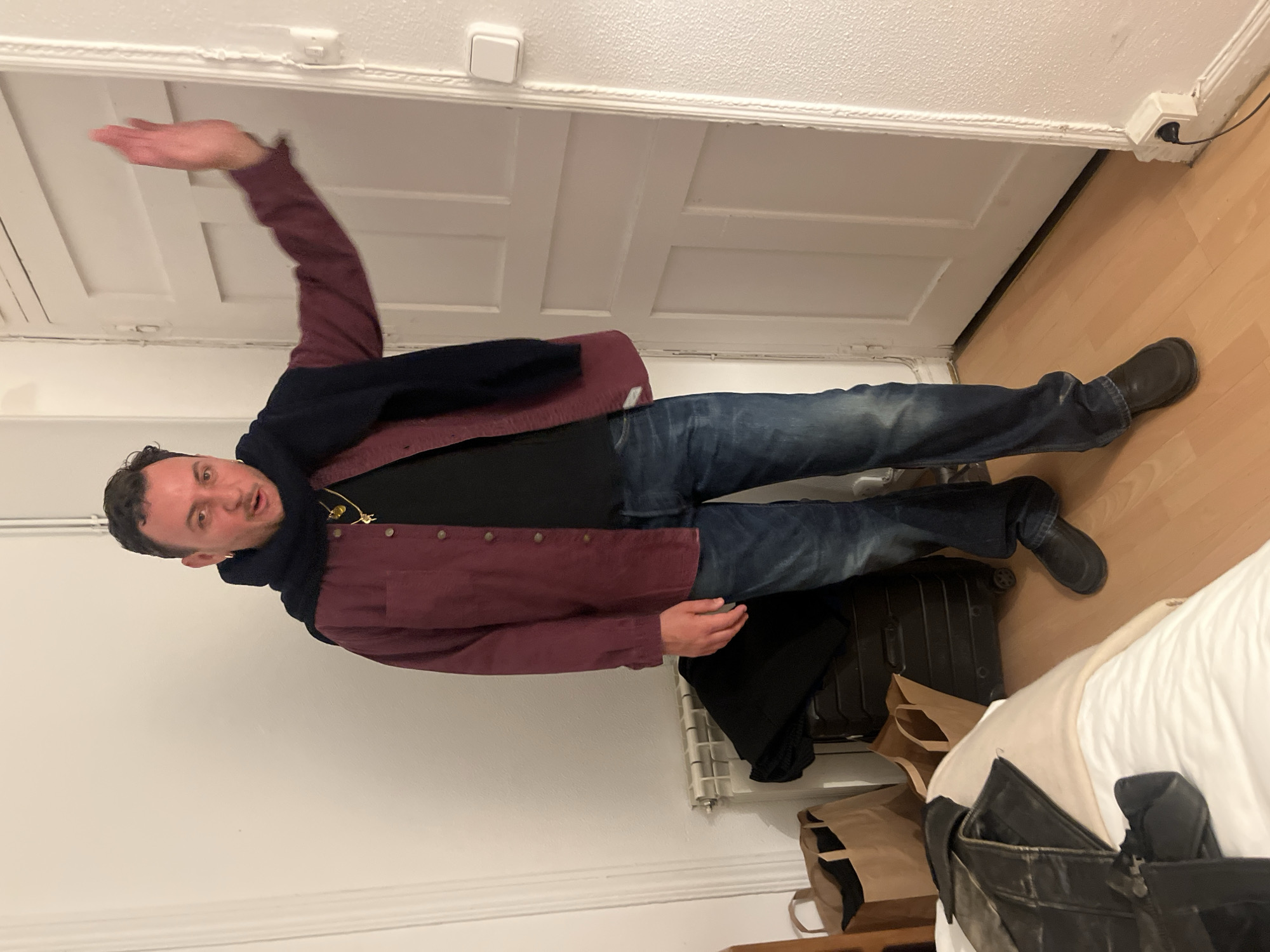 A man stands in a room. He wears jeans, black shoes, a black shirt, and a burgundy jacket, with a navy scarf draped around his neck. He has his right arm raised above his head. The room has a wooden floor and white walls, with a white door in the background. There is a suitcase, brown paper bags, and a black bag on the floor. The room is located in Madrid, Spain.
