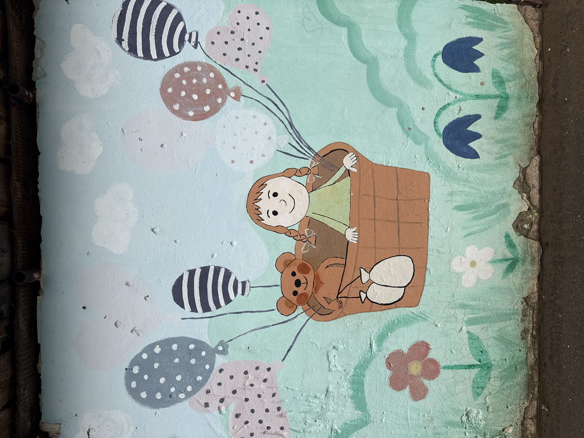 The image shows a section of an outdoor mural on a weathered wall in Votkinsk, Russia. The mural depicts a child with pigtails inside a brown basket, accompanied by a teddy bear. The basket is suspended by multiple balloons in varied patterns and colors. Additional painted details include flowers and stylized cloud shapes. The wall itself is cracked and has peeling paint, with an area of deterioration and exposed material along the right edge. Dark brown and gray materials, possibly wood and asphalt, are visible on the left and right edges of the image, respectively.