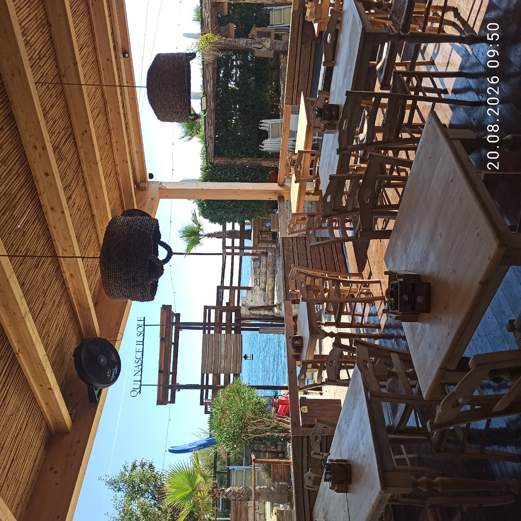 The image depicts an outdoor seating area of a restaurant or beach club, likely situated in the coastal region of Molfetta, Italy. The scene is viewed from a low angle, looking out towards the Adriatic Sea.
The foreground is dominated by wooden tables and chairs, arranged on a dark wooden floor that reflects sunlight. Some tables have wooden boxes with small plants or decorative items. The furniture appears rustic and casual, fitting for a seaside establishment.
In the midground, beyond the seating area, there is a sandy patch with some greenery, including palm trees. A blue paddleboard with "AQUATIC" written on it stands upright. Further back, a stone staircase leads up to a wooden deck with railings. The sea is visible as a calm, blue expanse.
Overhead, a thatched-like roof structure supported by wooden beams covers the seating area. Large, woven pendant lamps hang from the ceiling, and a black oscillating fan is mounted on one of the beams. There is a sign visible in the distance with text that appears to be "QUI NASCE IL SOLE".
The overall atmosphere is relaxed and summery, suggesting a place for dining and enjoying the coastal environment. The image is timestamped "20.08.2026 09:50". There are no people visible in the image.