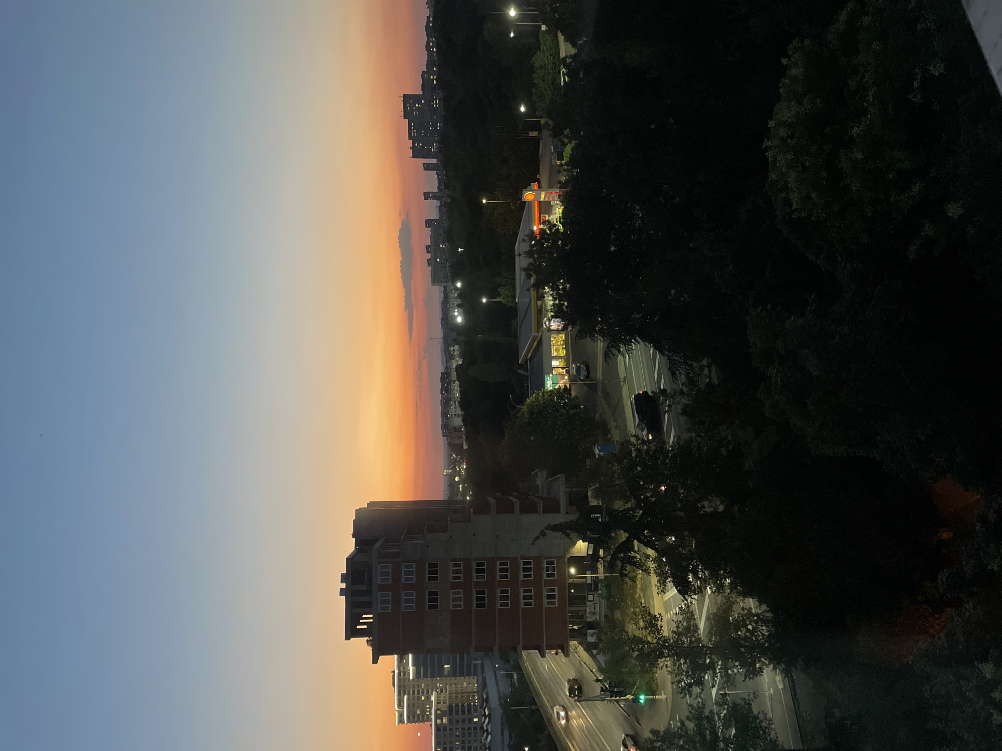 This is a high-angle, full shot of a city skyline at dusk. The sky transitions from a deep blue at the top to a vibrant orange and pink near the horizon, with a few wispy clouds visible. In the foreground, a dark silhouette of trees dominates the bottom portion of the image. To the left, a multi-story brick building, appearing to be under construction, stands tall with its windows reflecting the twilight. A road runs alongside this building, with a few cars visible. Further into the city, other buildings, including a modern high-rise with illuminated windows, are scattered amongst dark green trees. A gas station with illuminated signs is also visible in the middle ground. The overall scene depicts a residential and commercial area of a city during the transitional light of sunset.