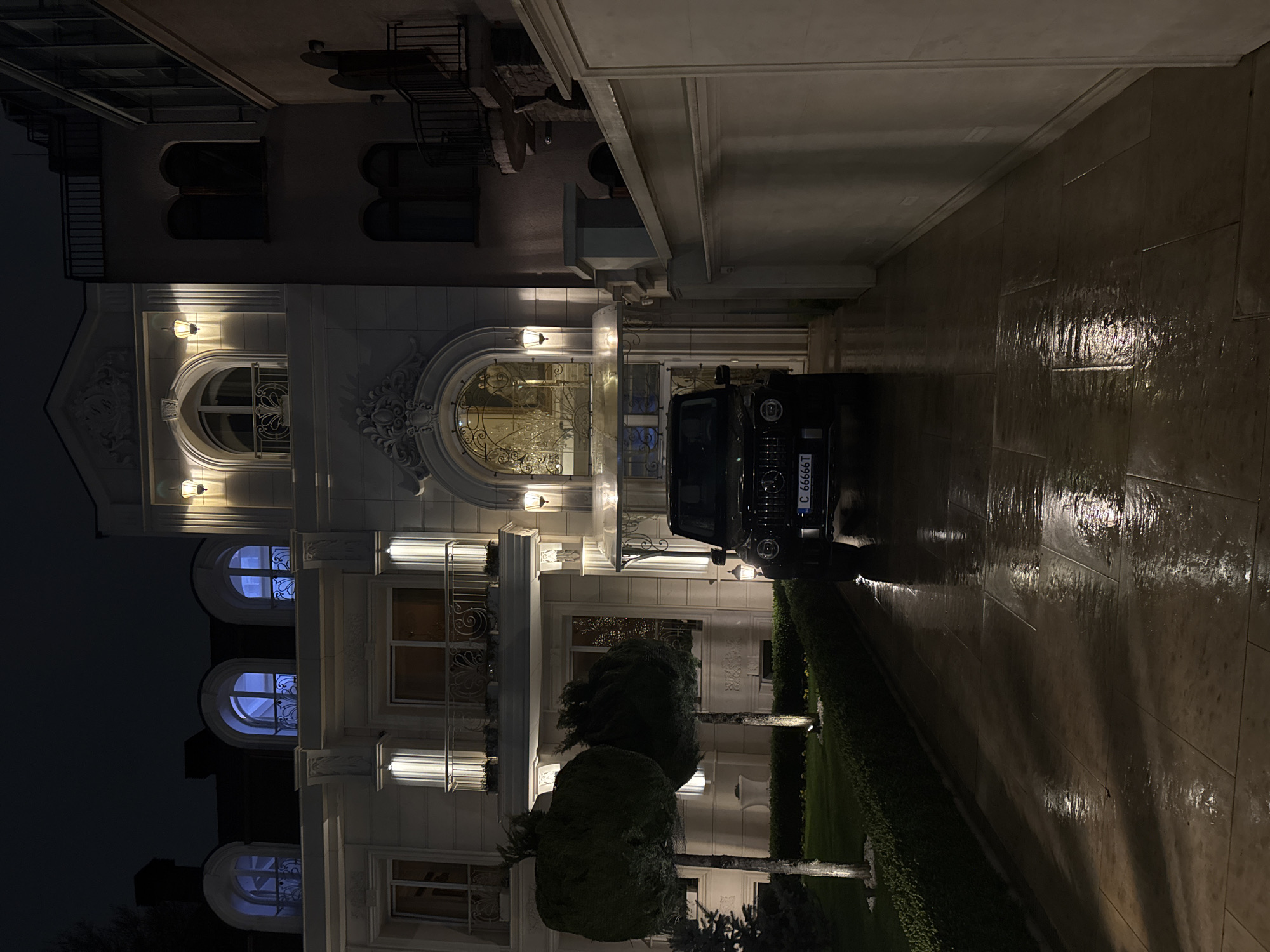 The image shows a black SUV parked in front of a large, ornately decorated building at night. The building appears to be a residence or a high-end establishment. The facade is illuminated with exterior lights, highlighting architectural details such as arched windows and decorative moldings. A wet, paved driveway leads to the building. The license plate of the vehicle indicates that the location is in Sofia, Bulgaria. The scene implies a wealthy or upscale neighborhood within the city. There are no people visible in the image.