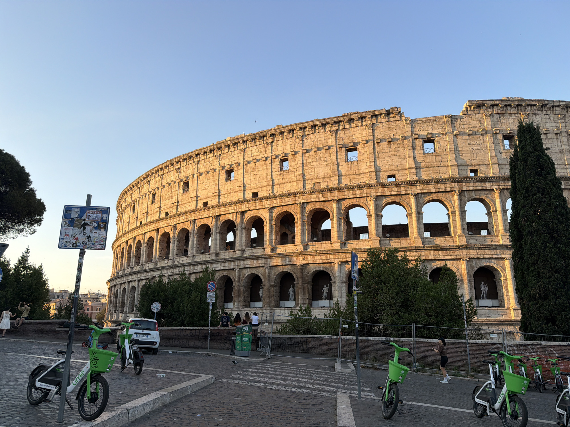 The image shows a wide view of the Colosseum in Rome, Italy, bathed in the warm light of a setting sun. In the foreground, several green electric scooters are parked on a cobbled street. A white car is visible behind a traffic sign. People are seen in the background, some sitting on a low wall, others walking. The Colosseum's iconic arches and weathered stone facade dominate the background, with lush green trees and a tall cypress tree framing the right side of the ancient structure. The sky is a clear, pale blue.